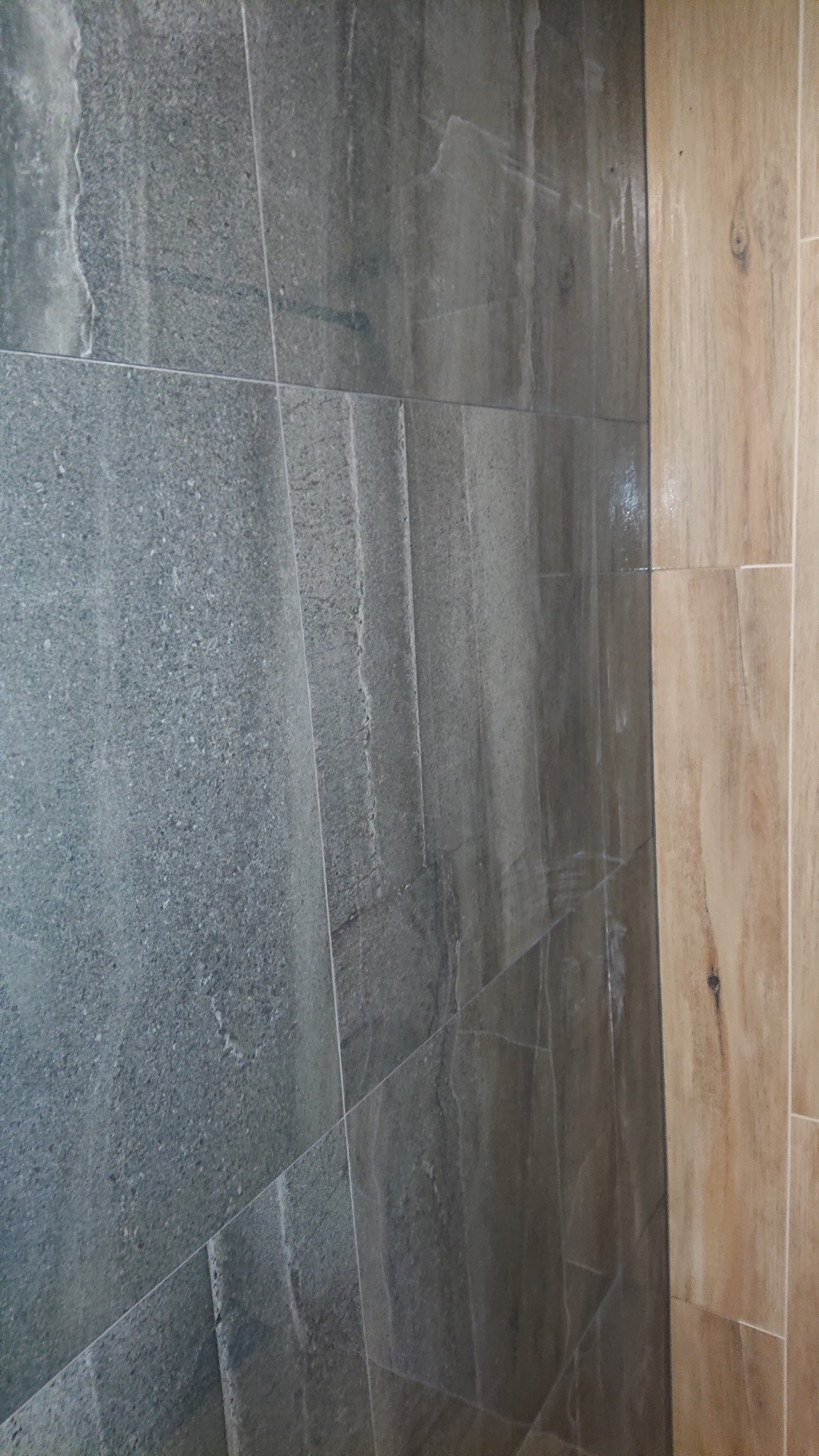 Ściana wyłożona szarymi płytkami imitującymi kamień oraz fragment ściany z jasnymi płytkami drewnopodobnymi, widoczne fugi.