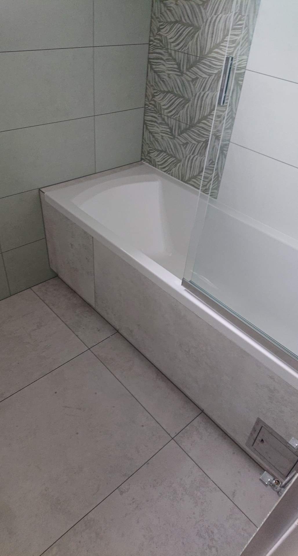 Wykończona łazienka: narożna wanna obudowana płytkami imitującymi beton, szklany parawan nawannowy, szare płytki podłogowe i ścienne, akcent z płytek dekoracyjnych z motywem liści.