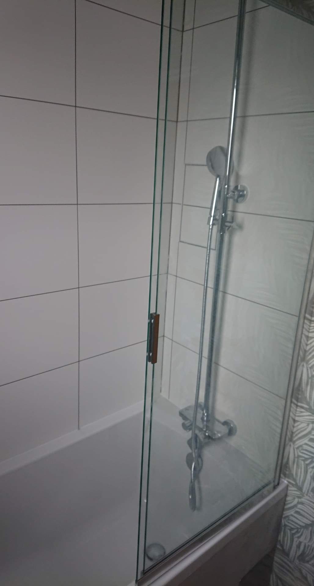 Kabina prysznicowa z chromowaną armaturą i białymi płytkami w łazience, widoczny fragment dekoracyjnej tapety w tle.