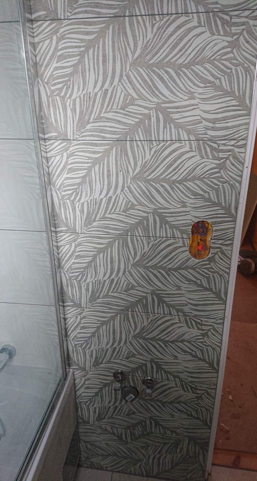 Ściana kabiny prysznicowej wyłożona płytkami z motywem liści palmowych, widoczne zawory i wnęka z przewodami elektrycznymi przed montażem armatury.