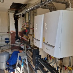 Hydro Craft - Instalacja dwóch białych pieców grzewczych De Dietrich z widocznymi rurami i zaworami w Małkini, instalator na drabinie w trakcie podłączania, widoczny fragment ściany z uszkodzonym tynkiem.