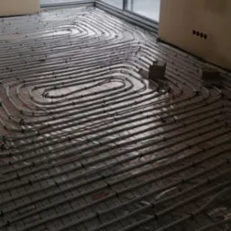 Białe rury ułożone spiralnie na podłodze, przygotowane do zalania wylewką, widoczne uchwyty mocujące i folia z nadrukiem, ściany w kolorze beżowym, okno zabezpieczone folią malarską.