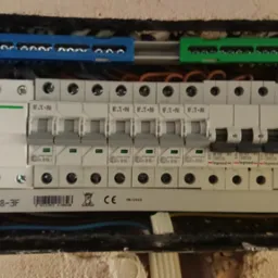 Rozdzielnica elektryczna z zainstalowanymi bezpiecznikami różnych marek (Schneider, Eaton, Legrand) oraz listwami zaciskowymi w kolorach niebieskim i zielonym, widoczne przewody.