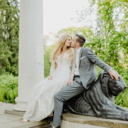 Karolina Berlińska - Para młoda: blondynka w koronkowej sukni i mężczyzna w szarym garniturze, całujący się siedząc na kamiennym lwie w parku, kolumna w tle.