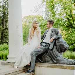 Karolina Berlińska - Para młoda w sesji plenerowej: panna młoda w koronkowej sukni z długim rękawem siedzi na schodach obok kamiennej rzeźby lwa, na której opiera się pan młody w szarym garniturze, w tle kolumny...