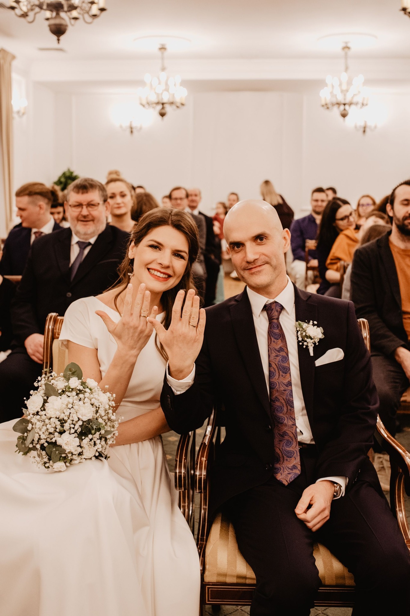 Para młoda siedzi w krzesłach, prezentując obrączki na tle gości w sali z żyrandolami.