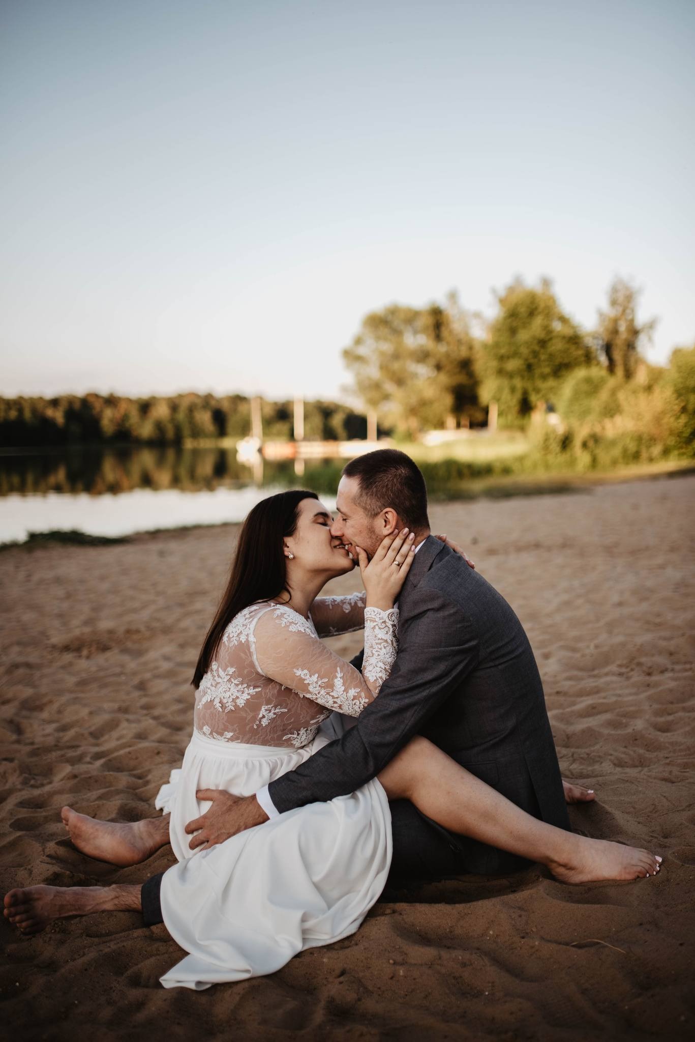 Para młoda siedzi na piasku nad jeziorem, całując się; Panna młoda w koronkowej sukni z długim rękawem, Pan młody w szarym garniturze.