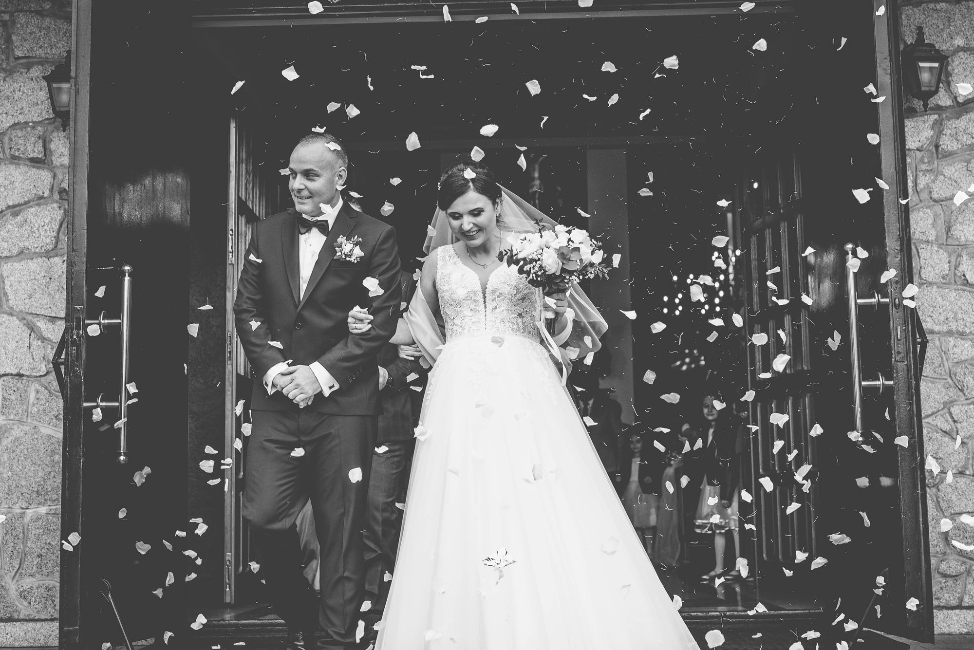 Czarno-białe zdjęcie: Nowożeńcy wychodzą z kościoła, obsypani płatkami róż, uśmiechnięta panna młoda trzyma bukiet.