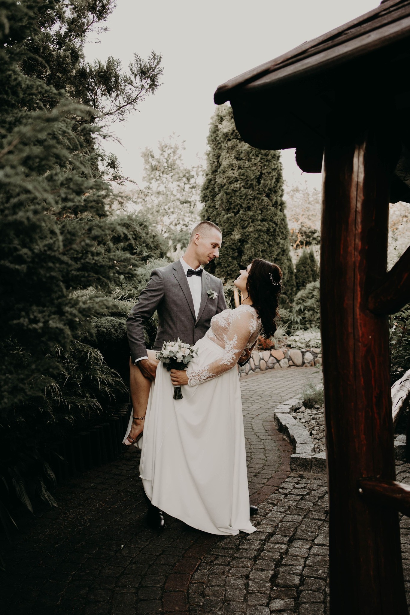 Pan młody trzyma pannę młodą na rękach w ogrodzie, panna młoda w białej sukni z koronkowymi rękawami, bukiet w dłoni, oboje patrzą na siebie z uśmiechem.