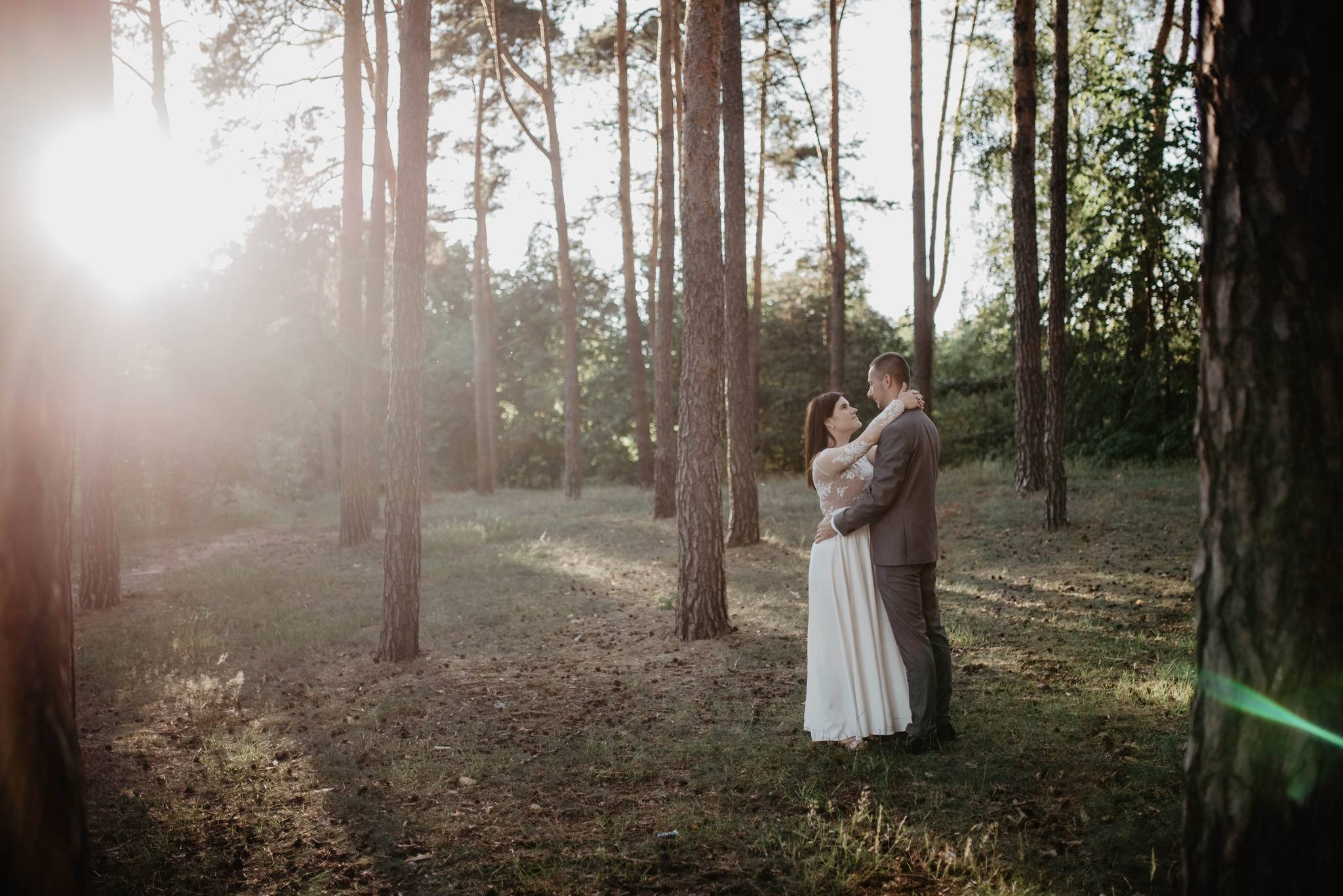 Para młoda obejmująca się w lesie sosnowym, panna młoda w sukni z koronkowymi rękawami, delikatne światło słoneczne przebija się przez drzewa.