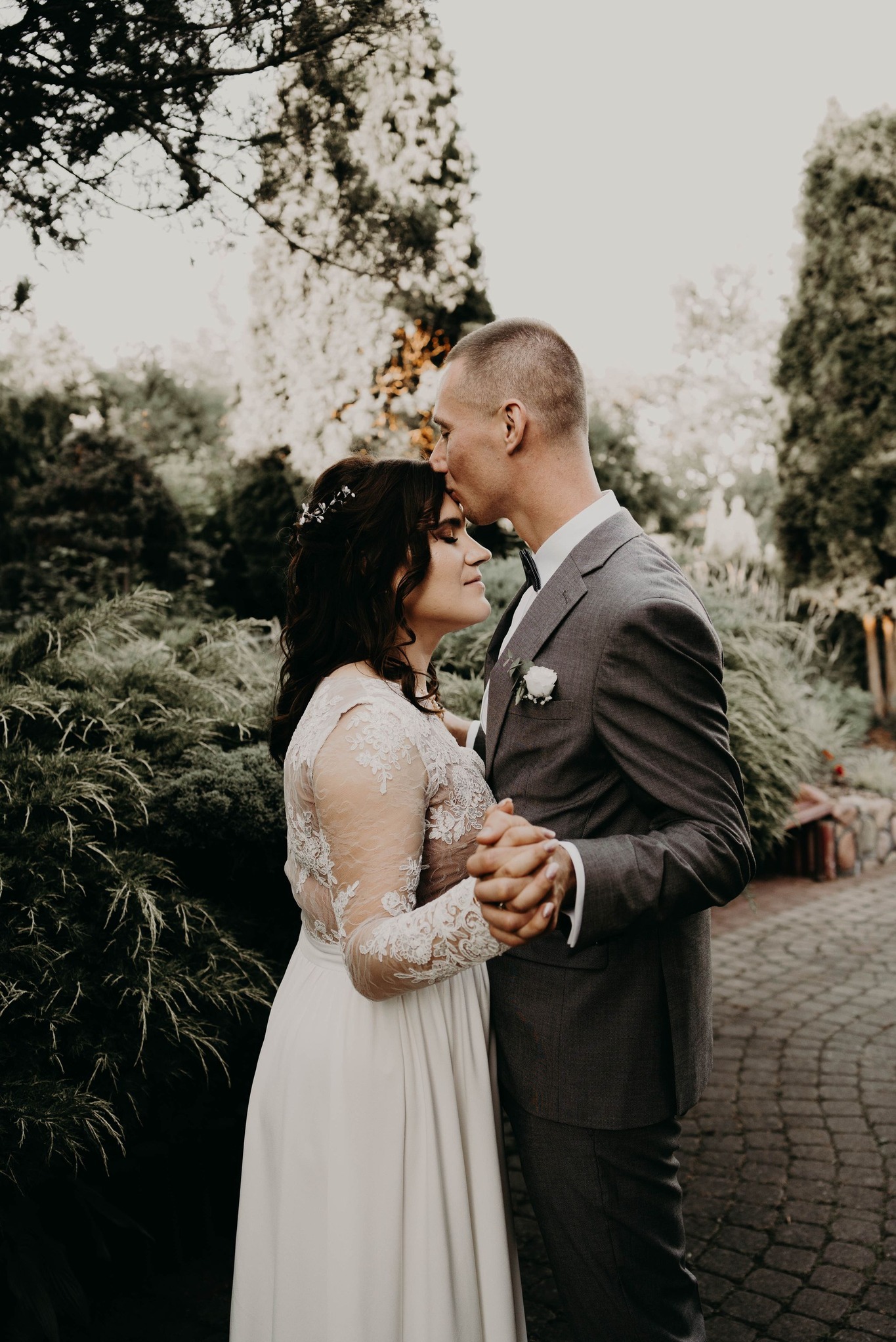 Elegancka para młoda w ogrodzie: pan młody całuje pannę młodą w czoło, trzymając ją za ręce, suknia z koronkowymi rękawami, szary garnitur.