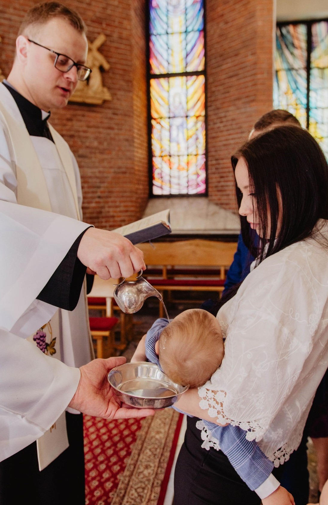 Ksiądz w okularach polewa wodą głowę niemowlęcia trzymanego przez matkę w kościele, w tle witrażowe okno.