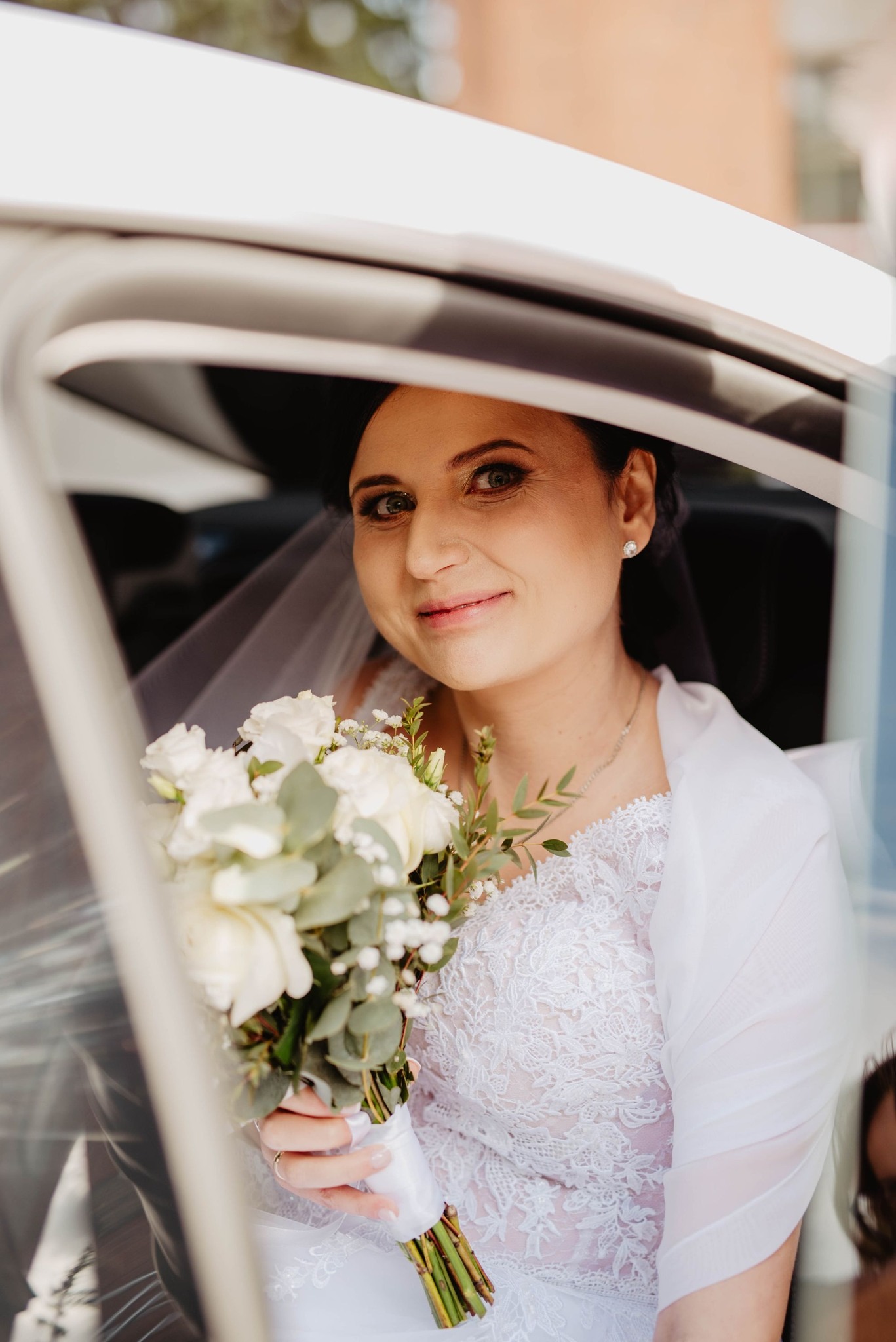 Panna młoda w białej sukni z koronką i bukietem białych róż, spogląda z uśmiechem z wnętrza białego samochodu, widoczny welon i kolczyk z brylantem.