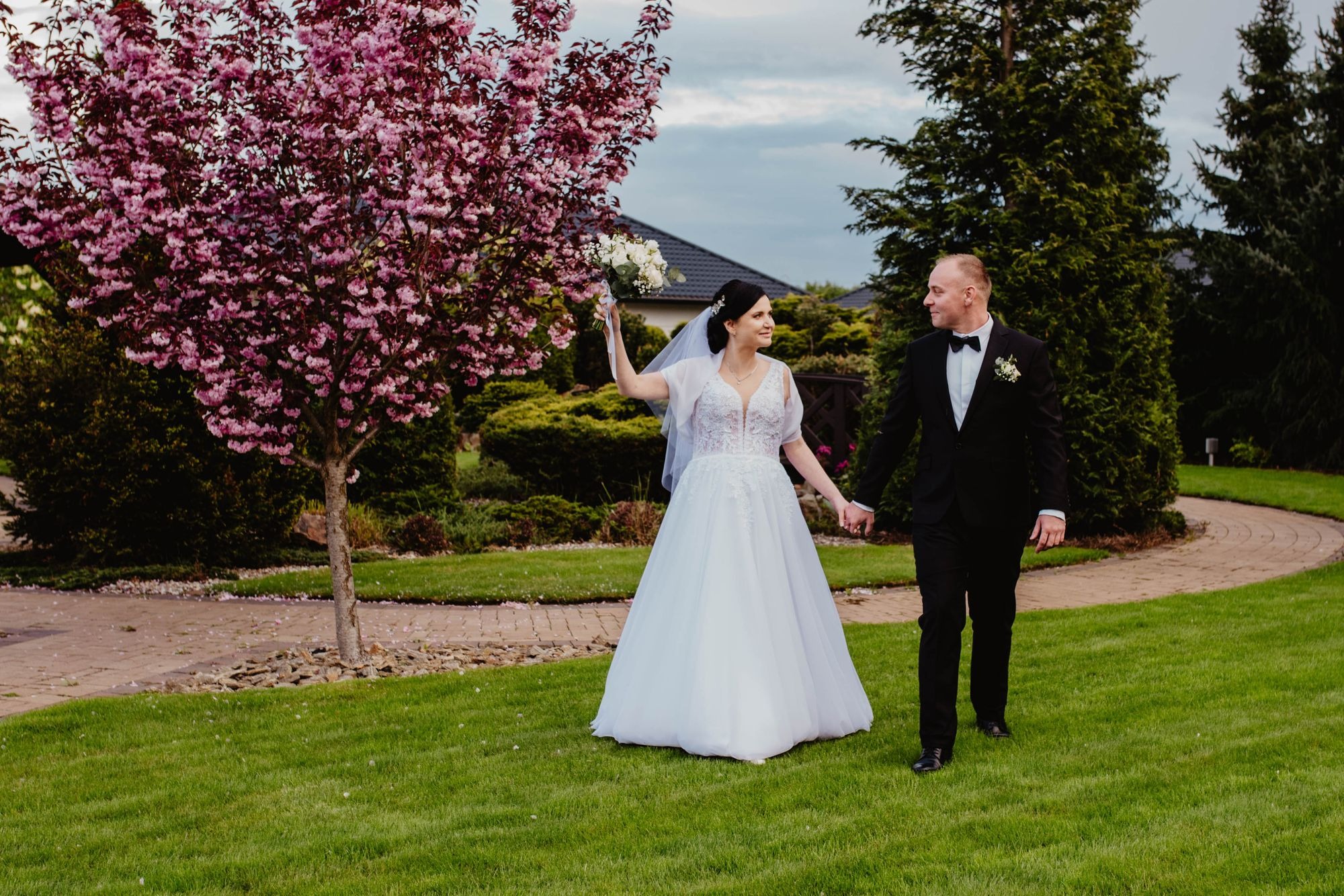 Para młoda spaceruje po trawniku, trzymając się za ręce, obok kwitnącego drzewa wiśniowego; panna młoda trzyma bukiet w górze.