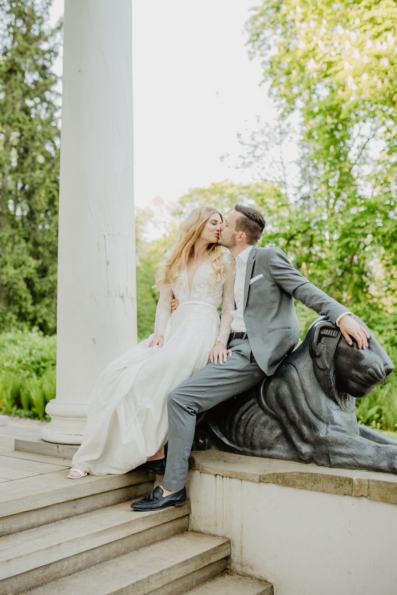 Para młoda: blondynka w koronkowej sukni i mężczyzna w szarym garniturze, całujący się siedząc na kamiennym lwie w parku, kolumna w tle.