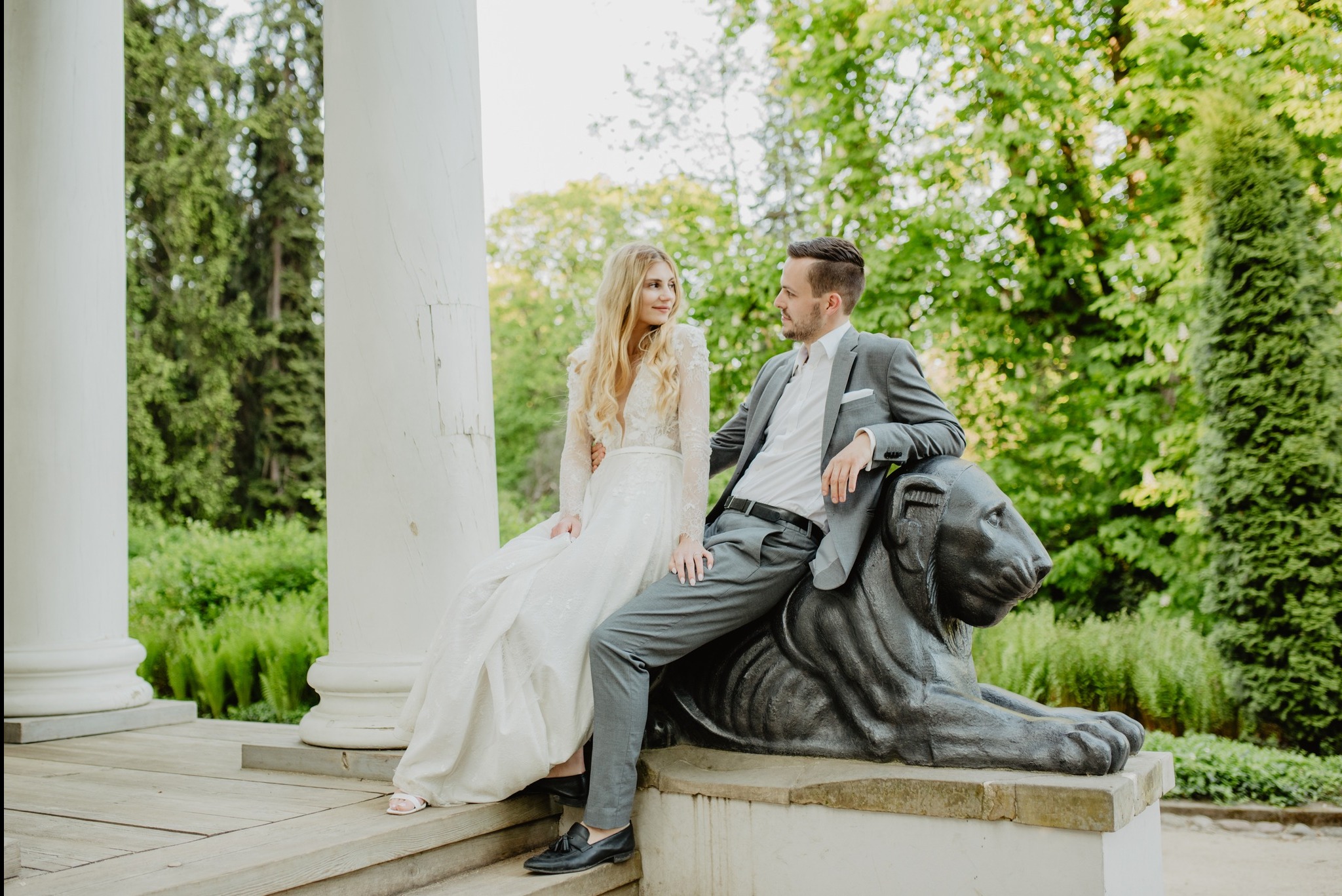 Para młoda w sesji plenerowej: panna młoda w koronkowej sukni z długim rękawem siedzi na schodach obok kamiennej rzeźby lwa, na której opiera się pan młody w szarym garniturze, w tle kolumny...