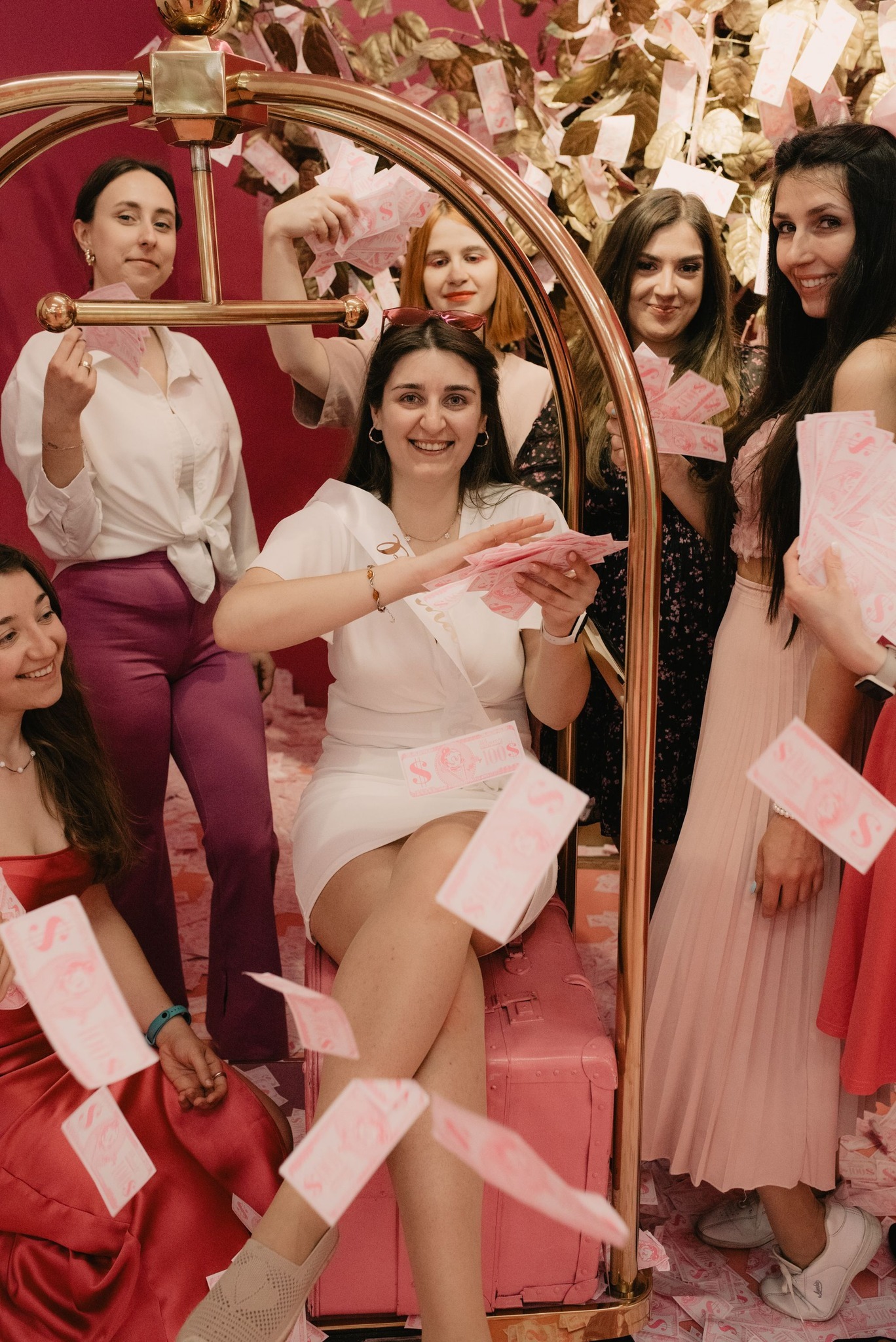 Grupa kobiet świętuje, rzucając w powietrze różowe banknoty, panna młoda siedzi na różowej walizce, otoczona przyjaciółkami w różnej kolorystyce ubrań, złoty wieszak na ubrania w tle, dekoracje...