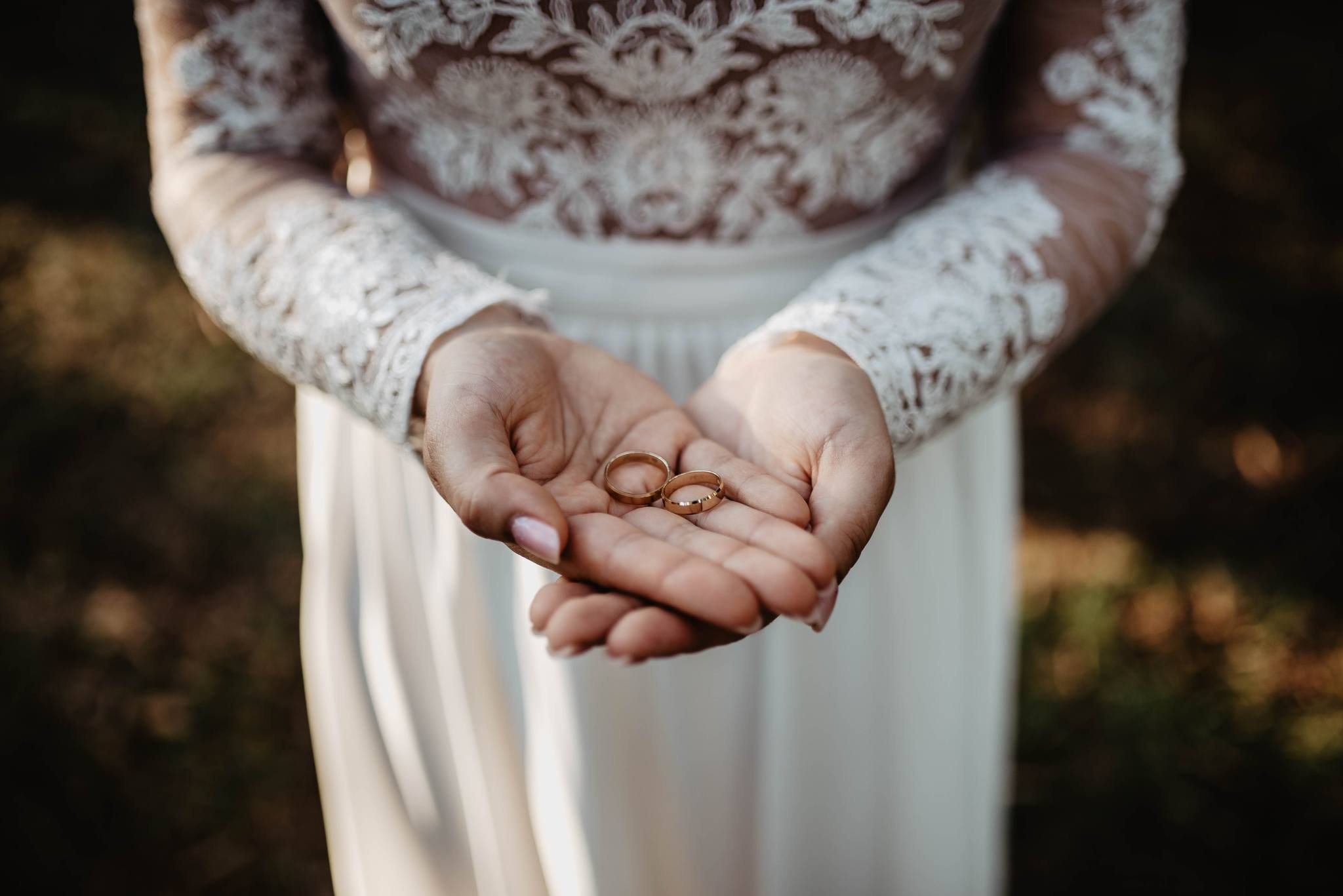 Dłonie panny młodej w koronkowej sukni trzymające dwie złote obrączki ślubne na tle rozmytego krajobrazu.