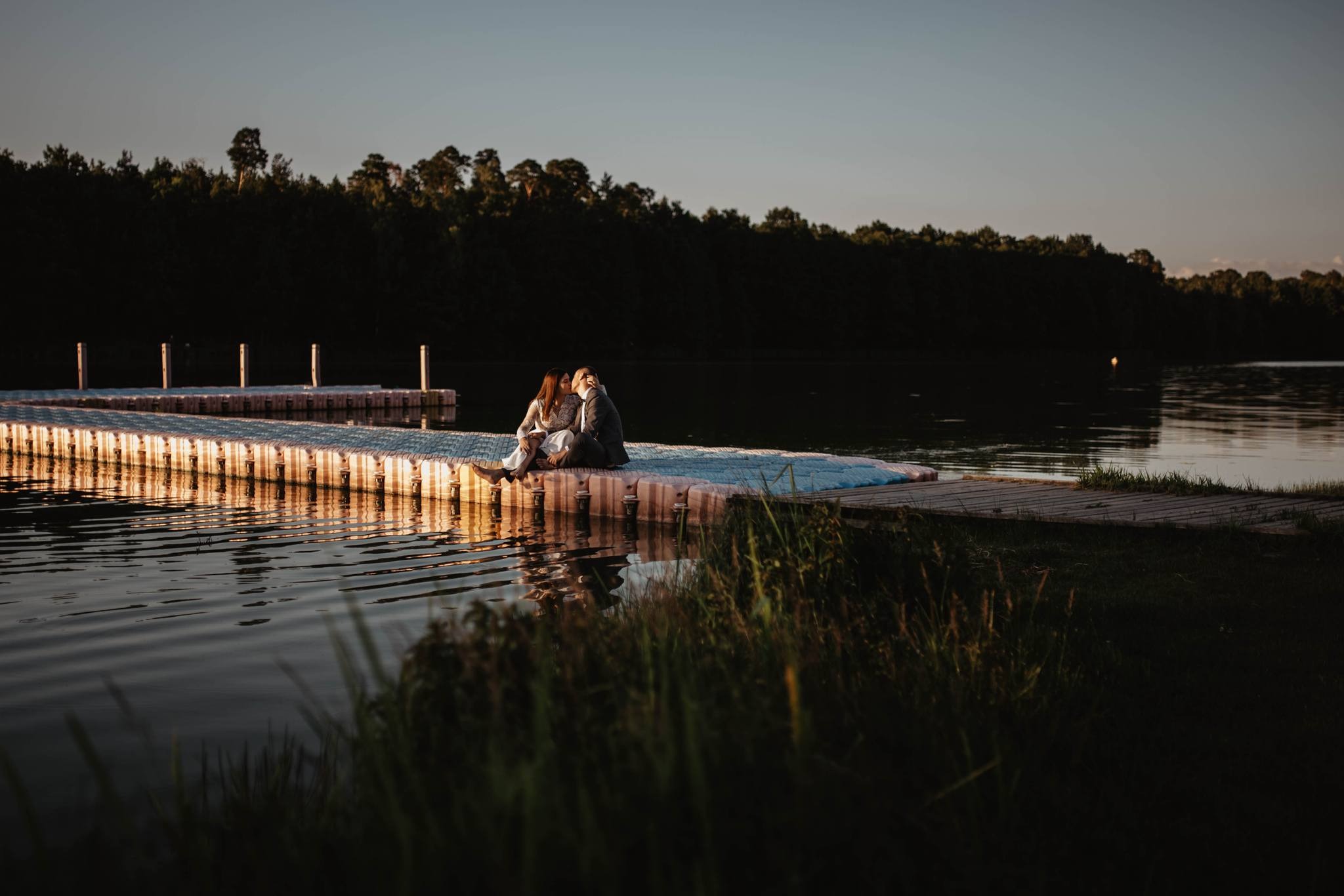 Para całująca się siedząc na pływającym pomoście nad jeziorem, w tle ciemny las i zachodzące słońce.