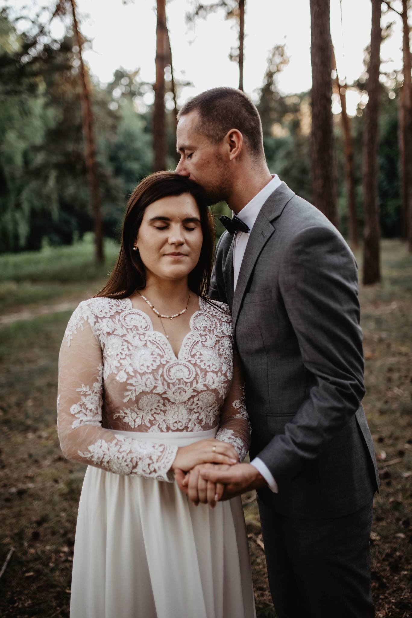 Para młoda w lesie: pan młody całuje pannę młodą w czoło, ona ma zamknięte oczy i trzyma go za rękę, na dłoni widoczny pierścionek