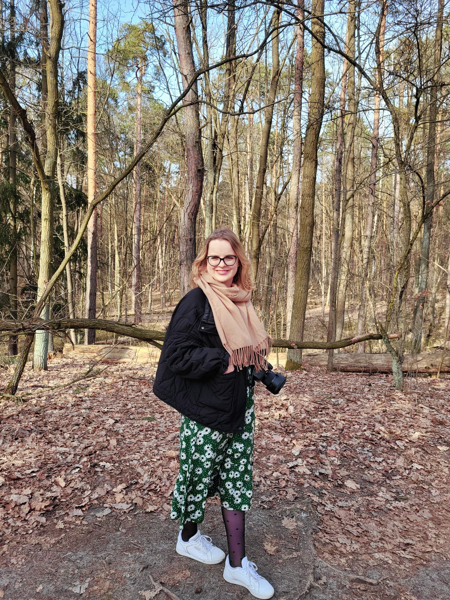 Kobieta w okularach, z aparatem w dłoni, pozuje w lesie w słoneczny dzień, ubrana w kwiecistą sukienkę i czarną kurtkę.