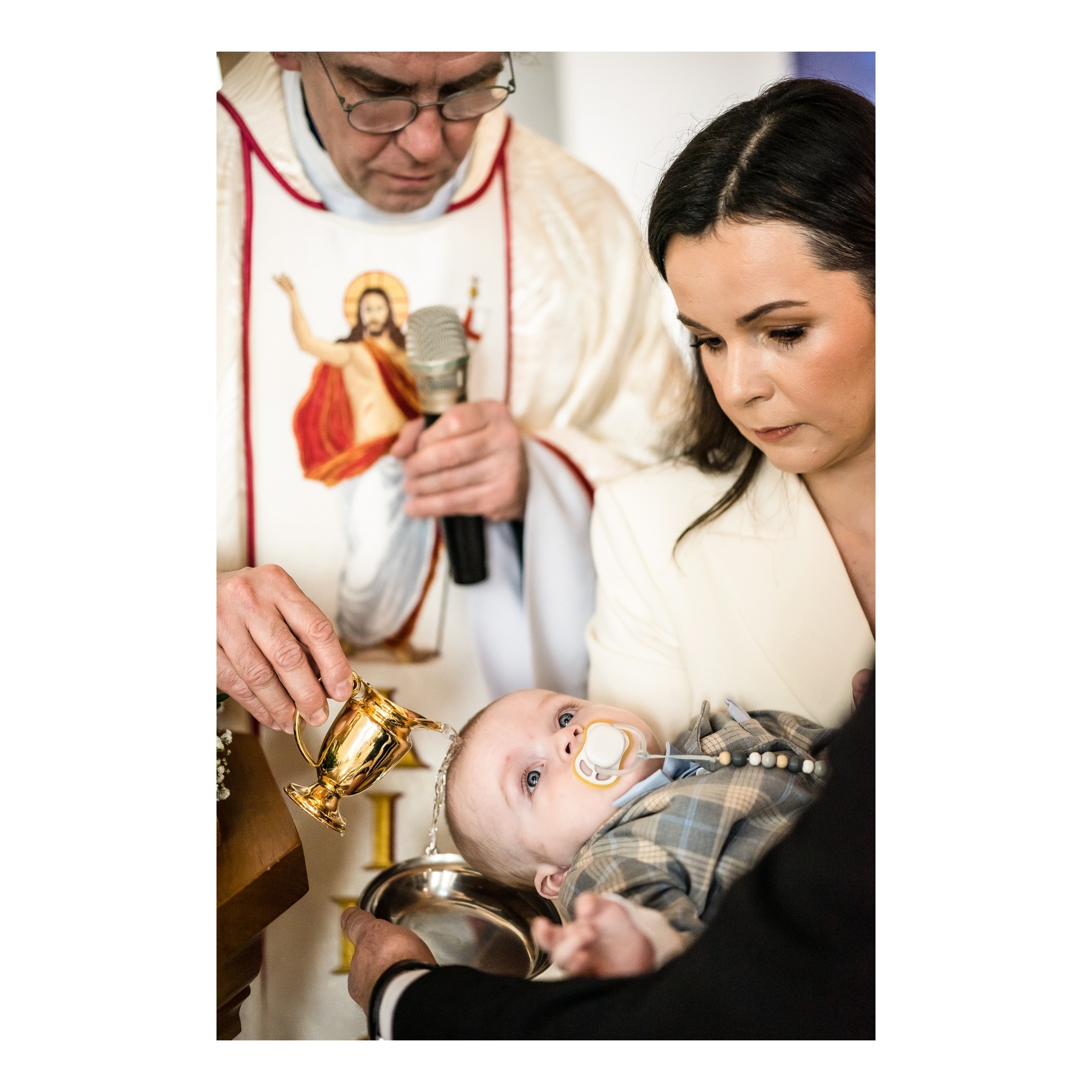 Ksiądz polewa wodą głowę niemowlęcia podczas chrztu, matka trzyma dziecko, w tle mikrofon i wizerunek Jezusa na szacie księdza.