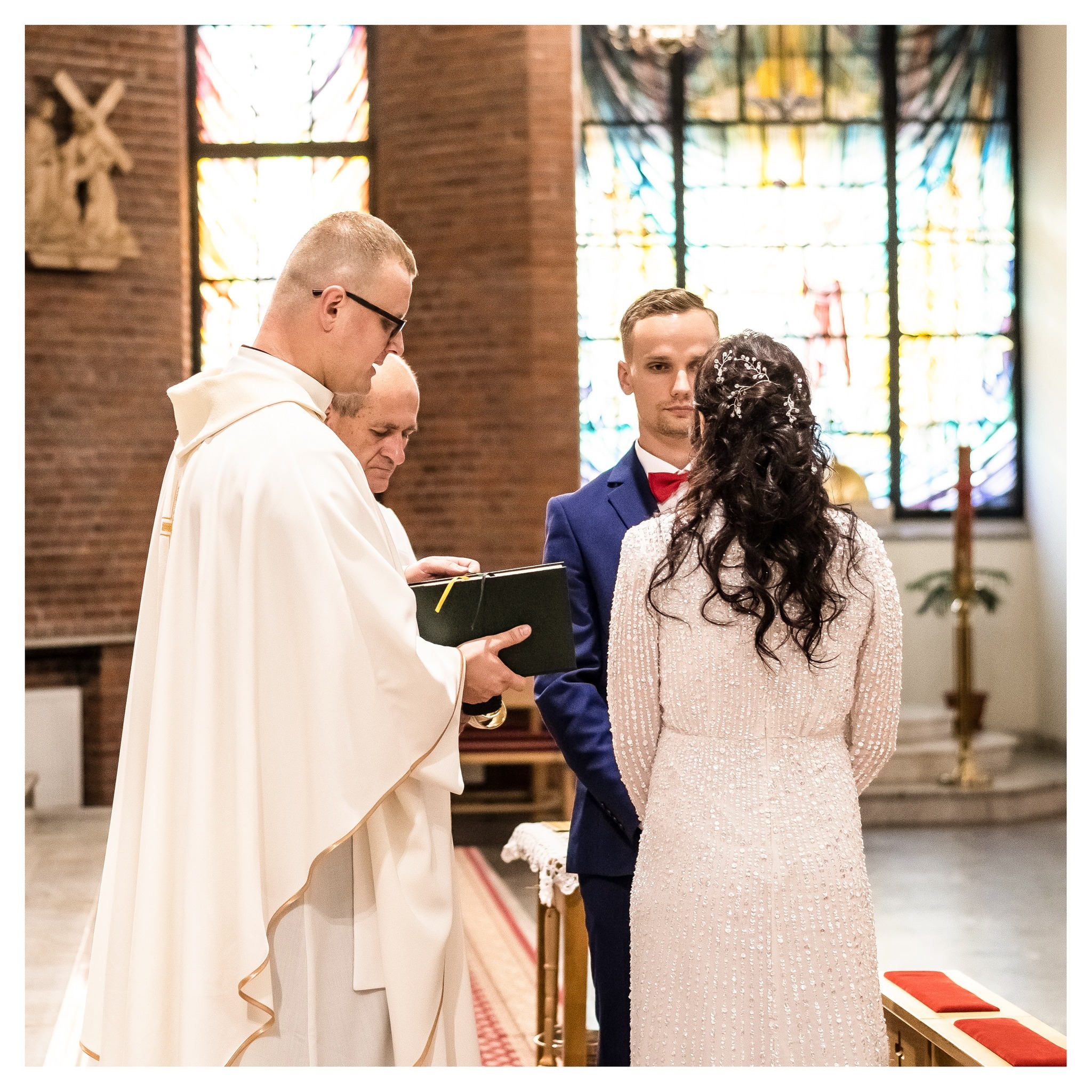 Para młoda podczas ceremonii ślubnej w kościele, ksiądz czyta z księgi, w tle witrażowe okno i rzeźba.