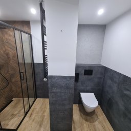 Brylant.BaU - Nowoczesna łazienka z kabiną prysznicową z geometrycznym wzorem na ścianie, wiszącą toaletą, grzejnikiem drabinkowym i podłogą imitującą drewno.