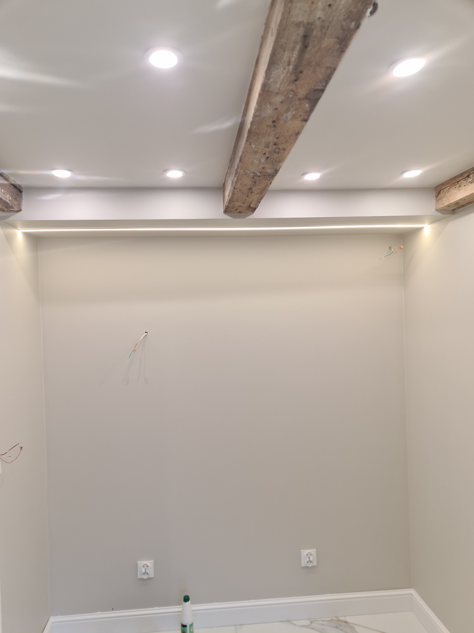 Wnętrze pokoju w trakcie wykańczania z drewnianymi belkami stropowymi, oświetleniem LED podsufitowym i wystającymi przewodami elektrycznymi ze ściany, widoczne gniazdka elektryczne i listwy...