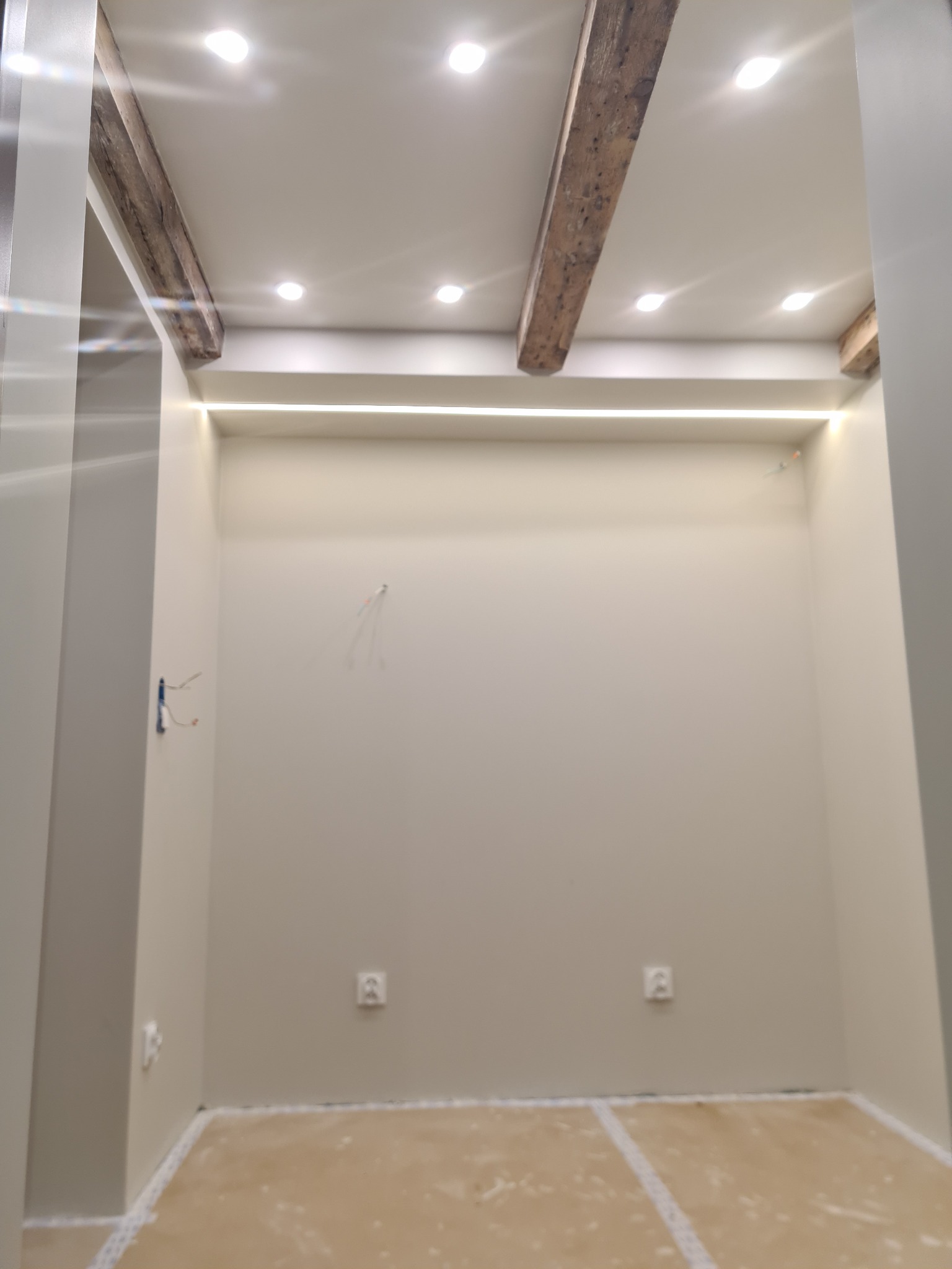 Pomalowane na beżowo wnętrze pokoju z odsłoniętymi drewnianymi belkami sufitowymi, oświetleniem punktowym i listwą LED pod sufitem, widoczne gniazdka elektryczne i wystające przewody.