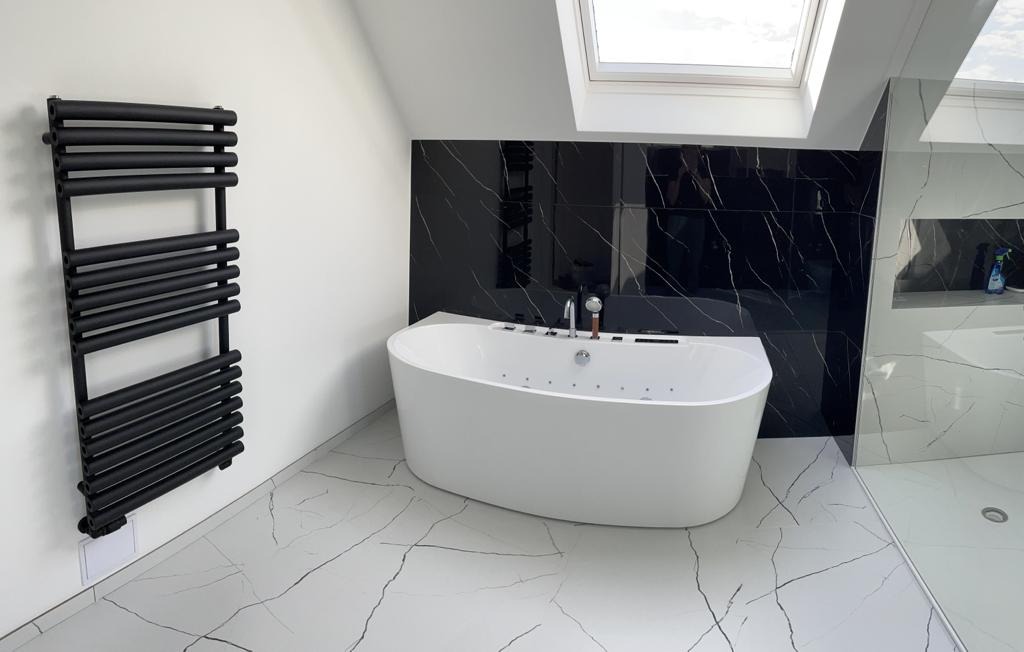 Nowoczesna łazienka na poddaszu z białą wanną z hydromasażem, czarnymi płytkami imitującymi marmur, grzejnikiem drabinkowym w kolorze czarnym i podłogą z białych płytek z czarnymi żyłami.
