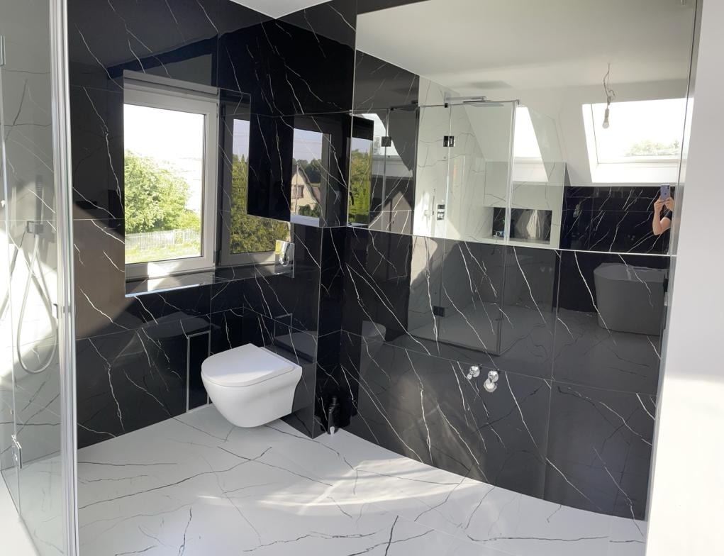 Nowoczesna łazienka z czarnymi płytkami imitującymi marmur z białymi żyłami, białymi płytkami podłogowymi z podobnym wzorem, kabiną prysznicową ze szkła, podwieszaną toaletą i dużym lustrem...