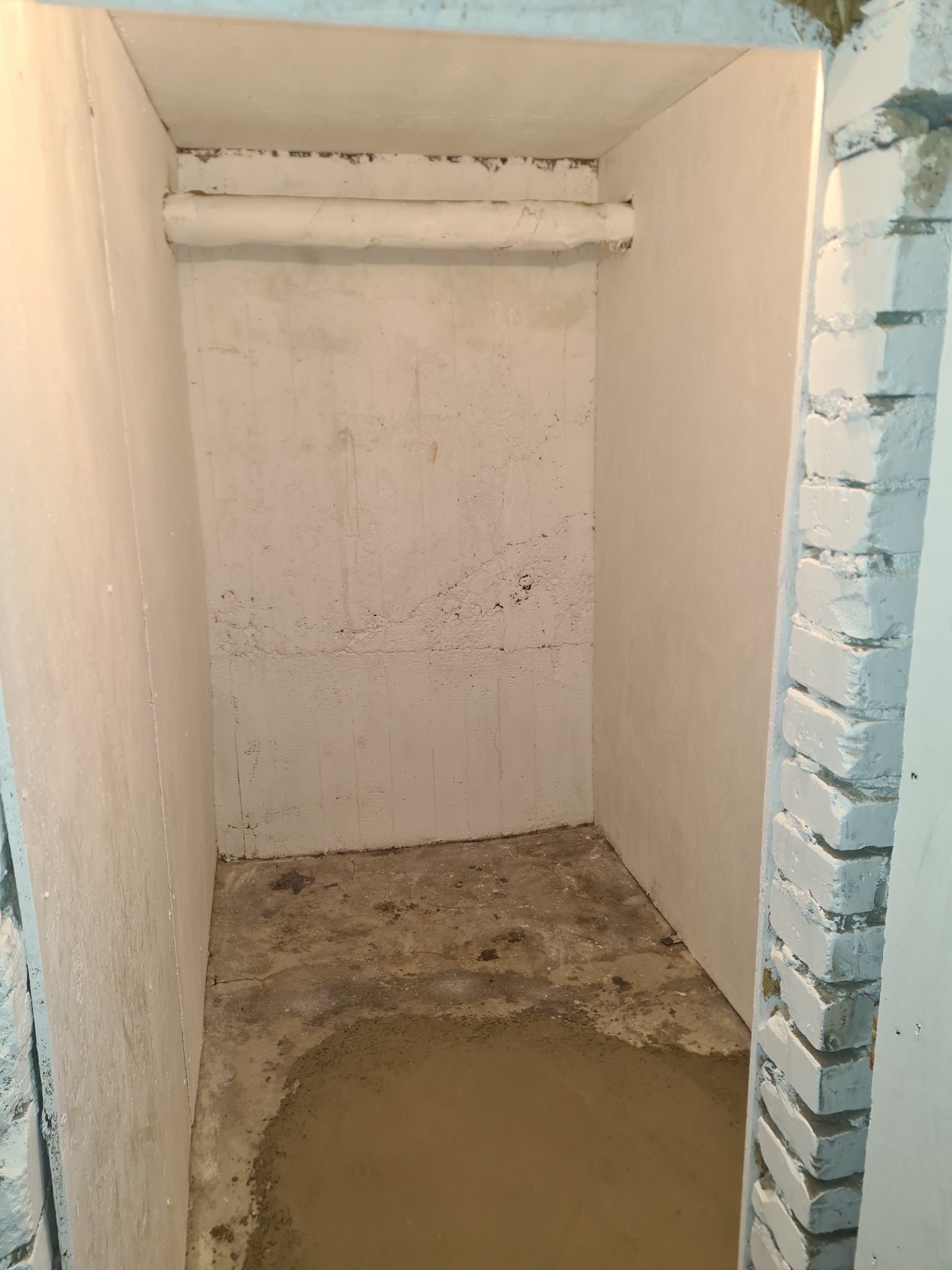 Wnętrze małego pomieszczenia w trakcie szpachlowania ścian, widoczne rury pod sufitem i fragment pomalowanej na biało ceglanej framugi drzwi.