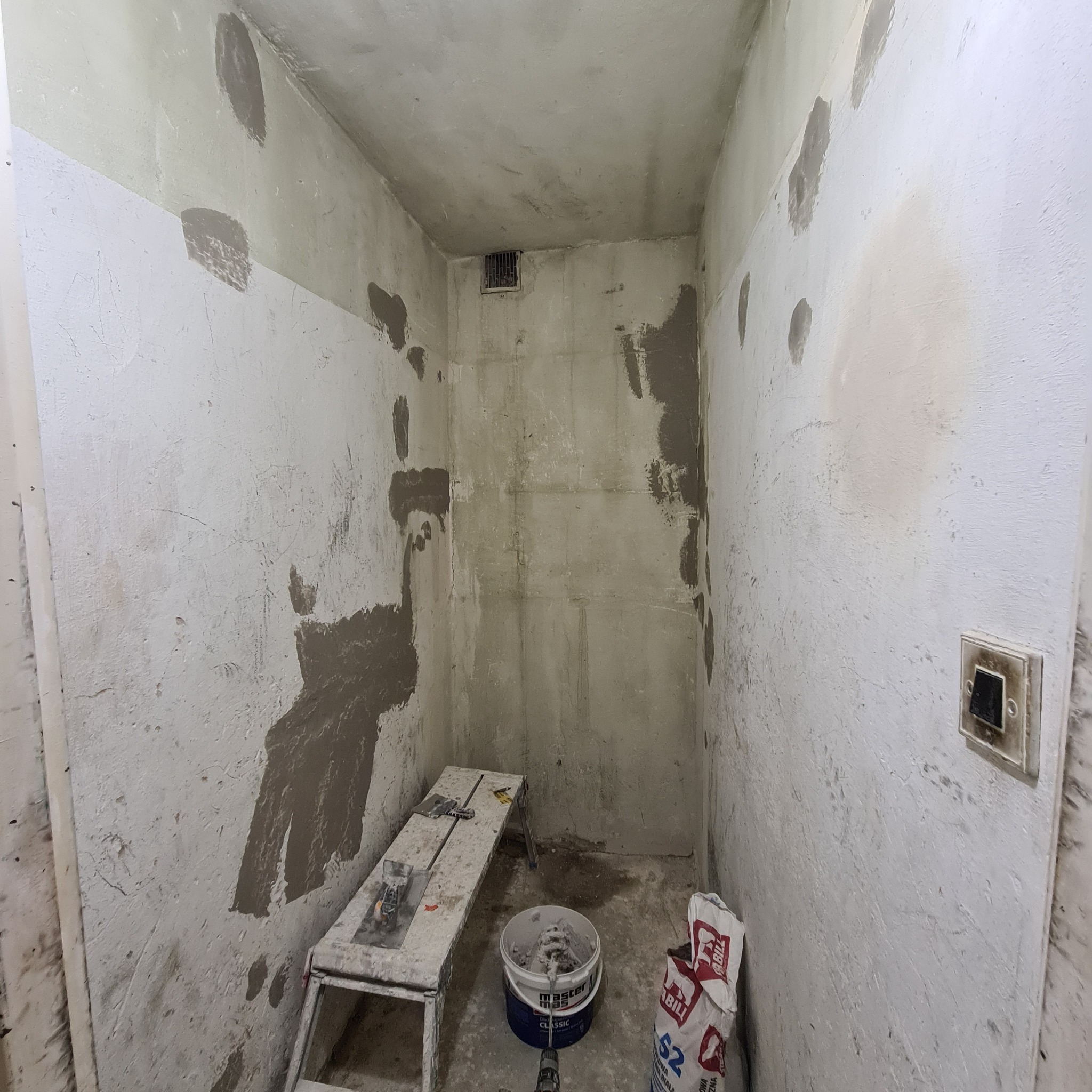 Narożne pomieszczenie w trakcie prac przygotowawczych do malowania, widoczne ślady po szpachlowaniu, drabina malarska, wiadro z zaprawą i worek gładzi szpachlowej.