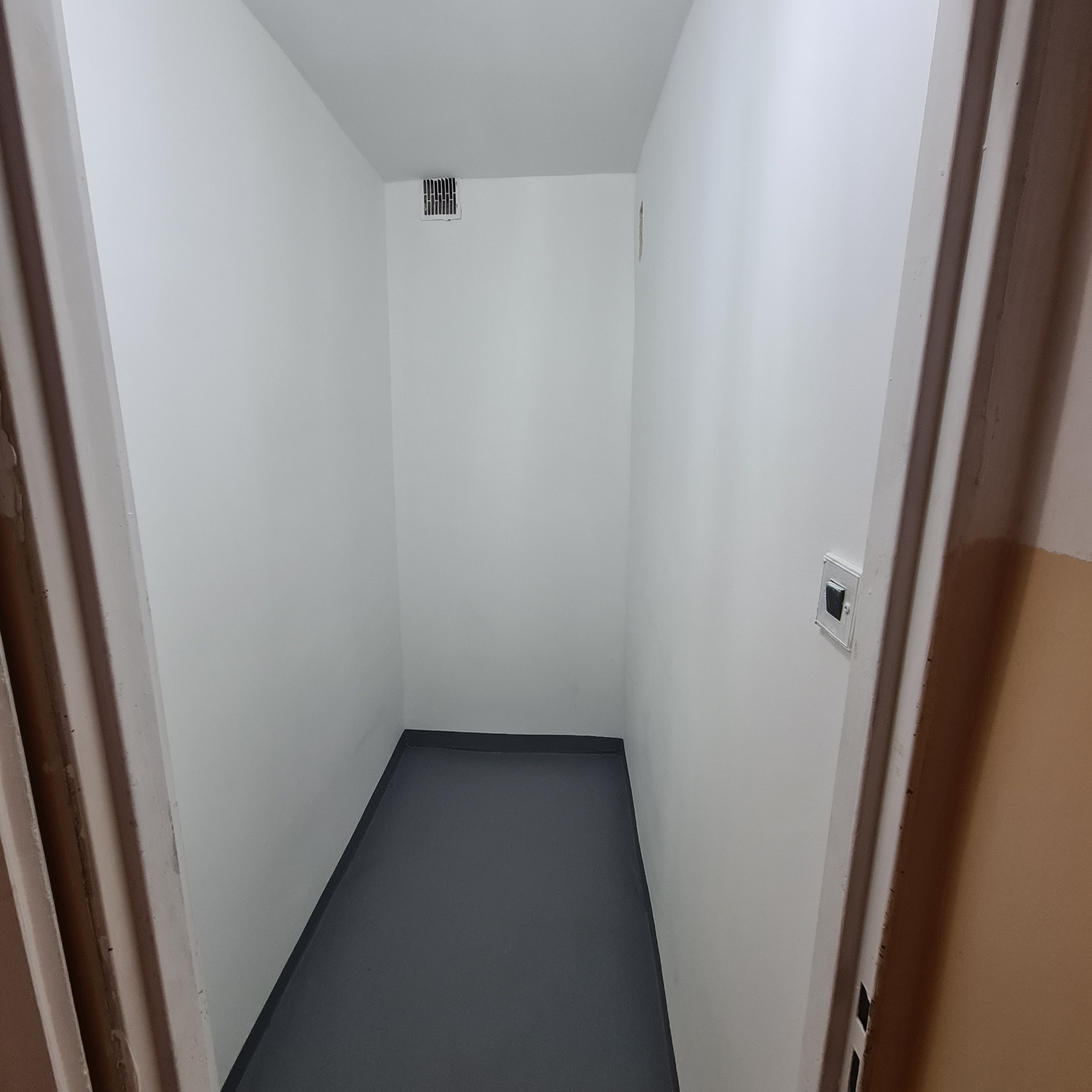 Wnętrze małego pomieszczenia po odświeżeniu: świeżo pomalowane białe ściany, szara podłoga, widoczna kratka wentylacyjna pod sufitem i włącznik światła na ścianie, widoczne fragmenty ościeżnicy.