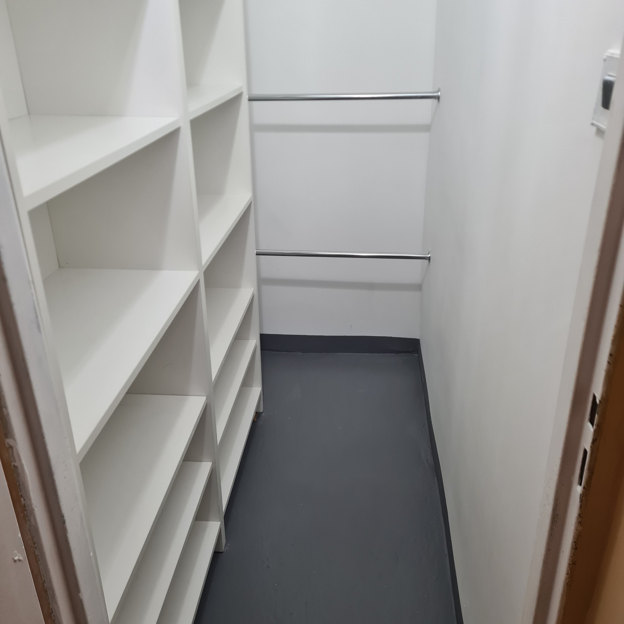 Wnętrze garderoby z białymi półkami i dwoma drążkami na ubrania, szara podłoga, widok z perspektywy wejścia.
