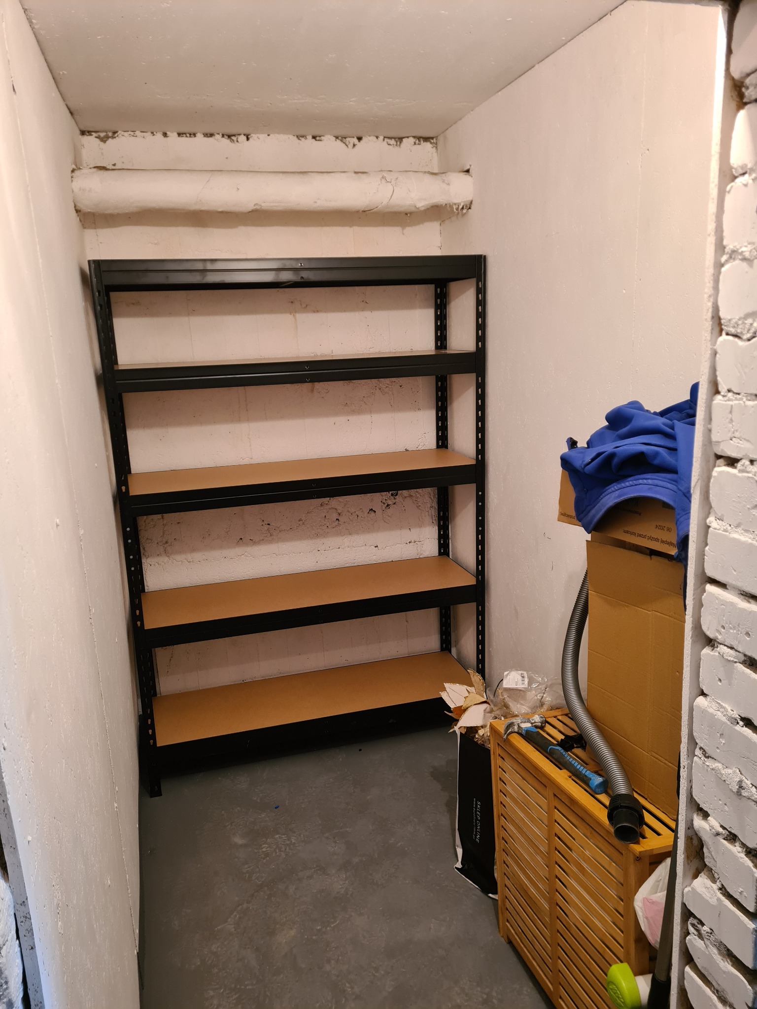 Półki metalowe z drewnopodobnymi blatami w trakcie montażu w pomieszczeniu gospodarczym w Ełku, widoczne surowe ściany i rury pod sufitem, elementy wyposażenia obok.