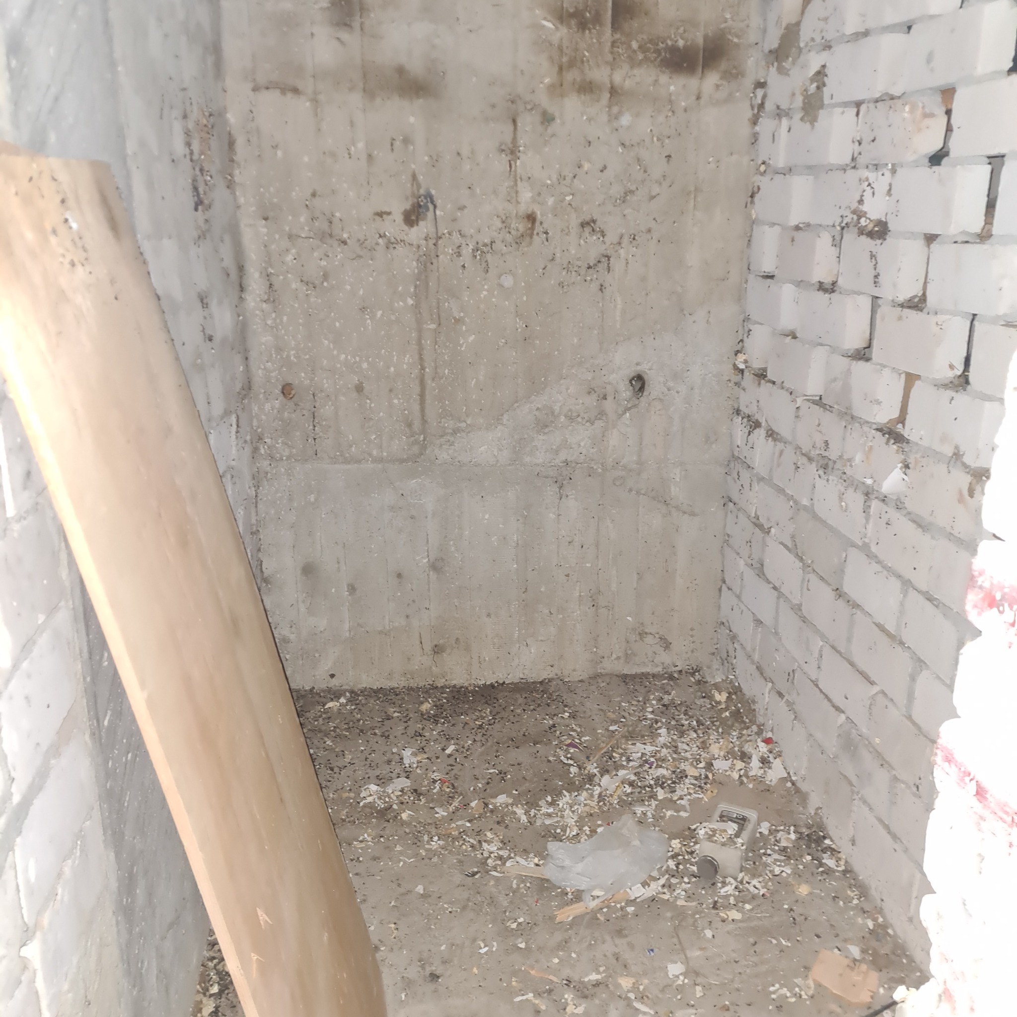 Surowe wnętrze w trakcie prac wykończeniowych: betonowa ściana z widocznymi śladami po instalacjach, fragment ściany z białej cegły, na podłodze resztki materiałów budowlanych, oparta deska.