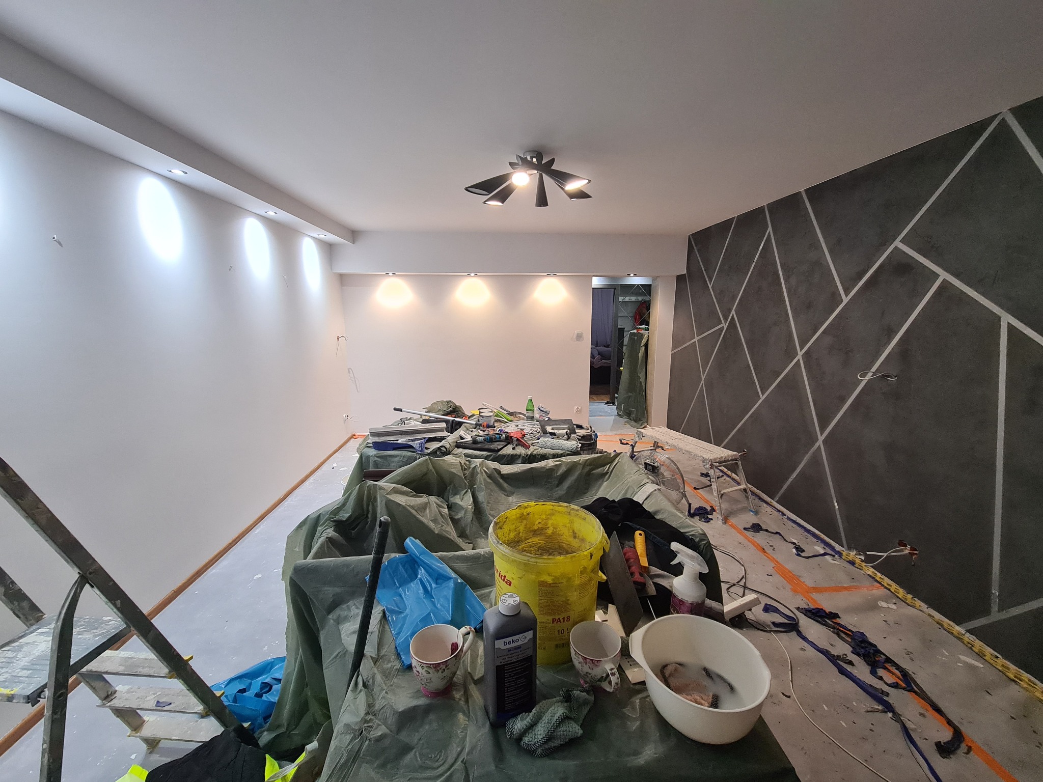 Pokój w trakcie odświeżania: pomalowana na biało ściana z oświetleniem punktowym, druga ściana z dekoracyjnym wzorem geometrycznym, podłoga zabezpieczona folią, na środku rozłożone narzędzia...