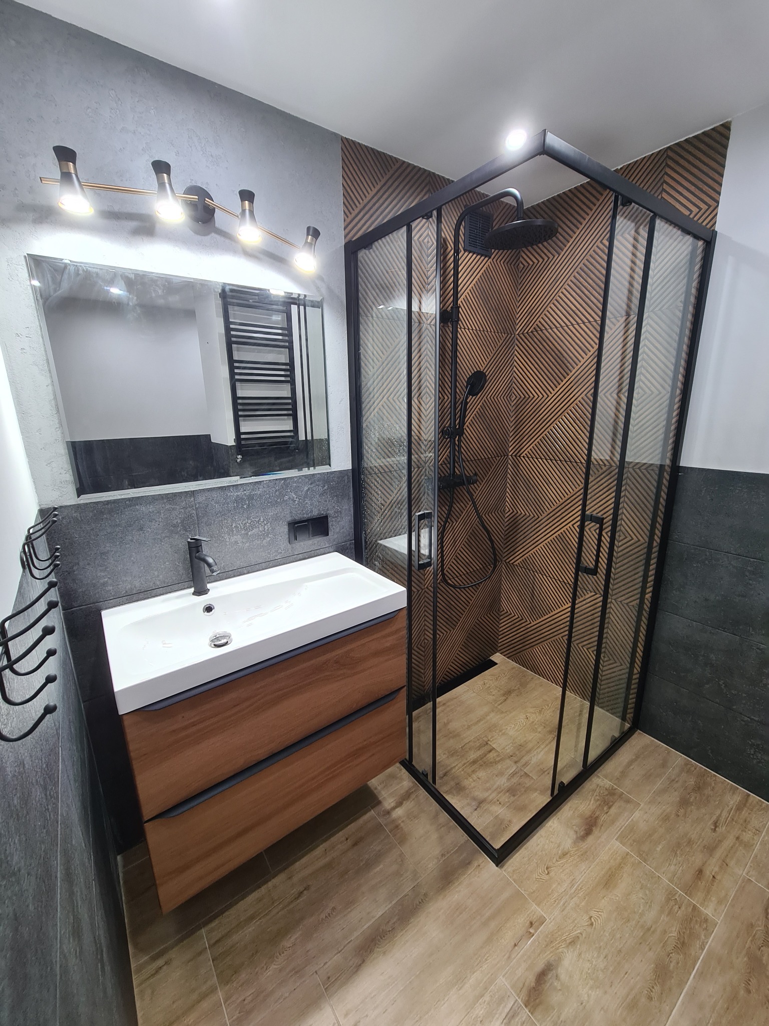 Nowoczesna łazienka z drewnianą szafką pod umywalką, czarną armaturą i kabiną prysznicową z geometryczną ścianą z płytek w odcieniach brązu.