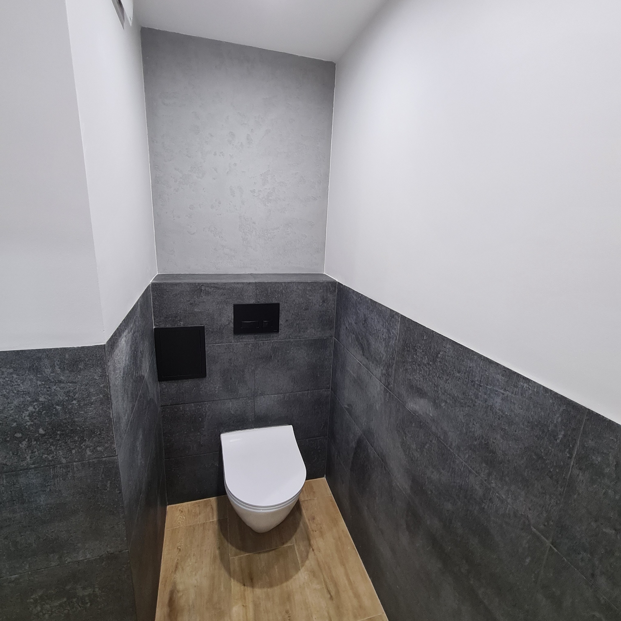 Nowoczesna toaleta w małej łazience z szarą, teksturowaną ścianą i płytkami imitującymi kamień, drewnopodobna podłoga, wisząca muszla klozetowa z czarnym panelem sterującym.