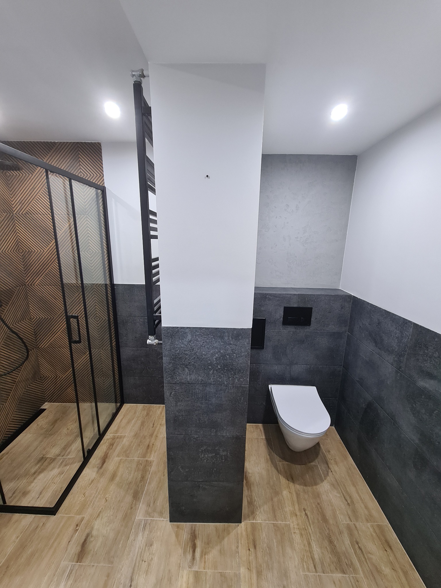 Nowoczesna łazienka z kabiną prysznicową z geometrycznym wzorem na ścianie, wiszącą toaletą, grzejnikiem drabinkowym i podłogą imitującą drewno.