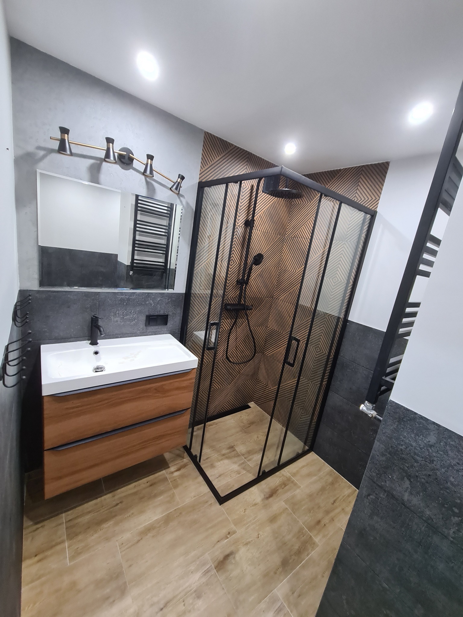 Nowoczesna łazienka z kabiną prysznicową w czarnej ramie, drewnianą szafką pod umywalką i geometryczną mozaiką na ścianie.