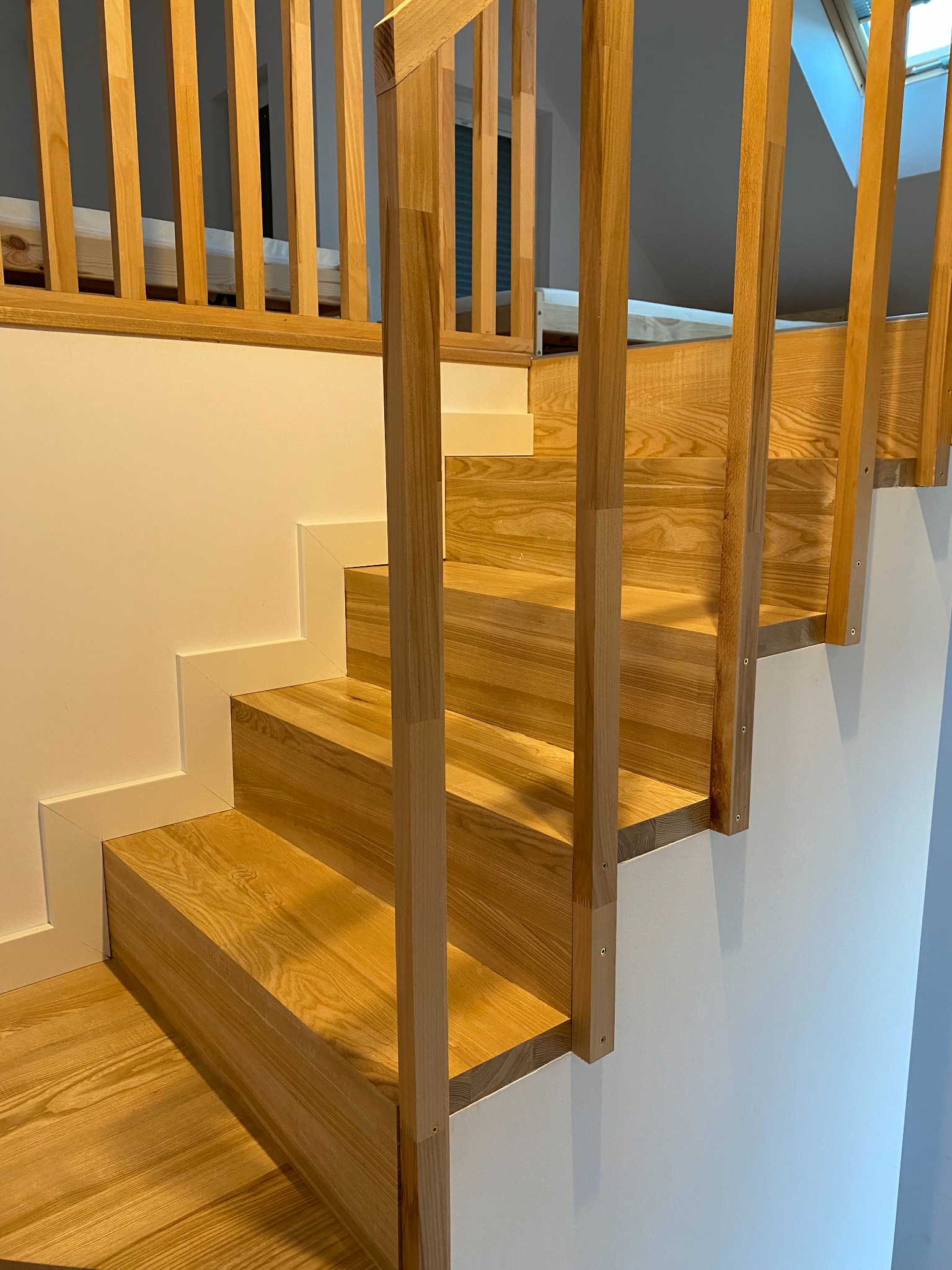 Jasne, drewniane schody z balustradą, widoczne mocowania słupków do stopni, białe wykończenie ścian.