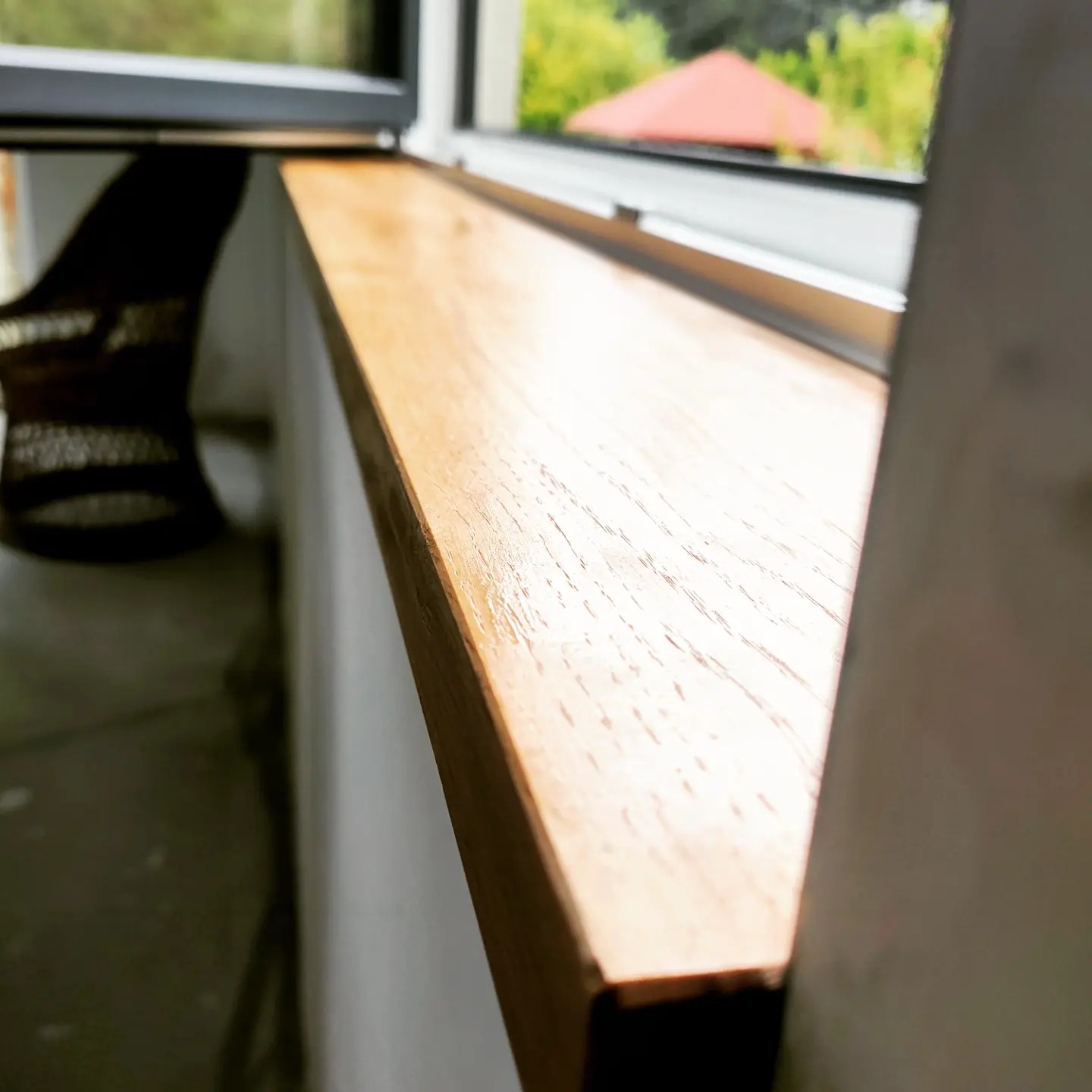 Drewniany parapet okienny z widoczną strukturą drewna, świeżo polakierowany, z widokiem na zieleń i fragment dachu za oknem.