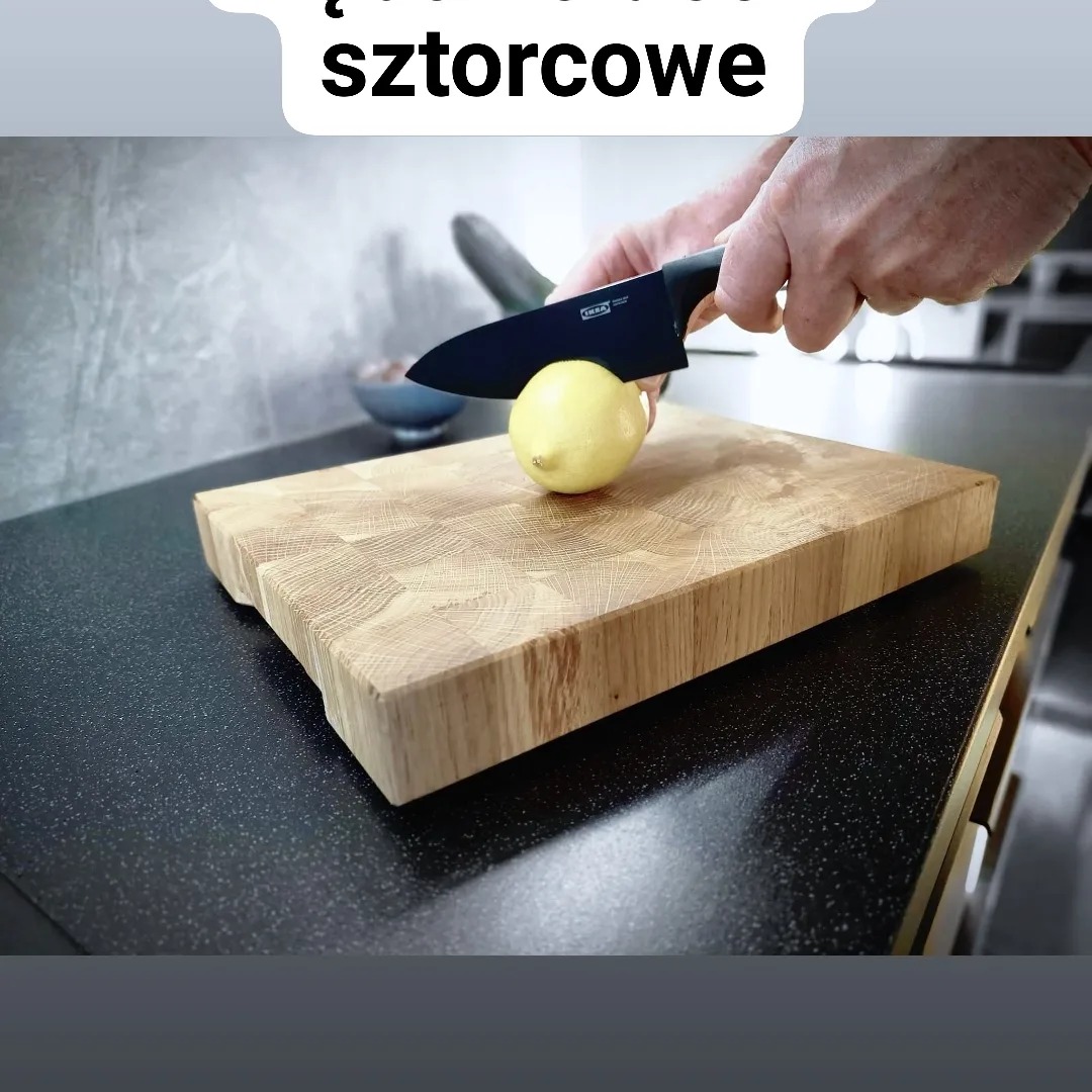 Ręka kroi cytrynę na grubej, drewnianej desce do krojenia z widocznym usłojeniem, nóż z czarnym ostrzem, czarny blat kuchenny w tle.
