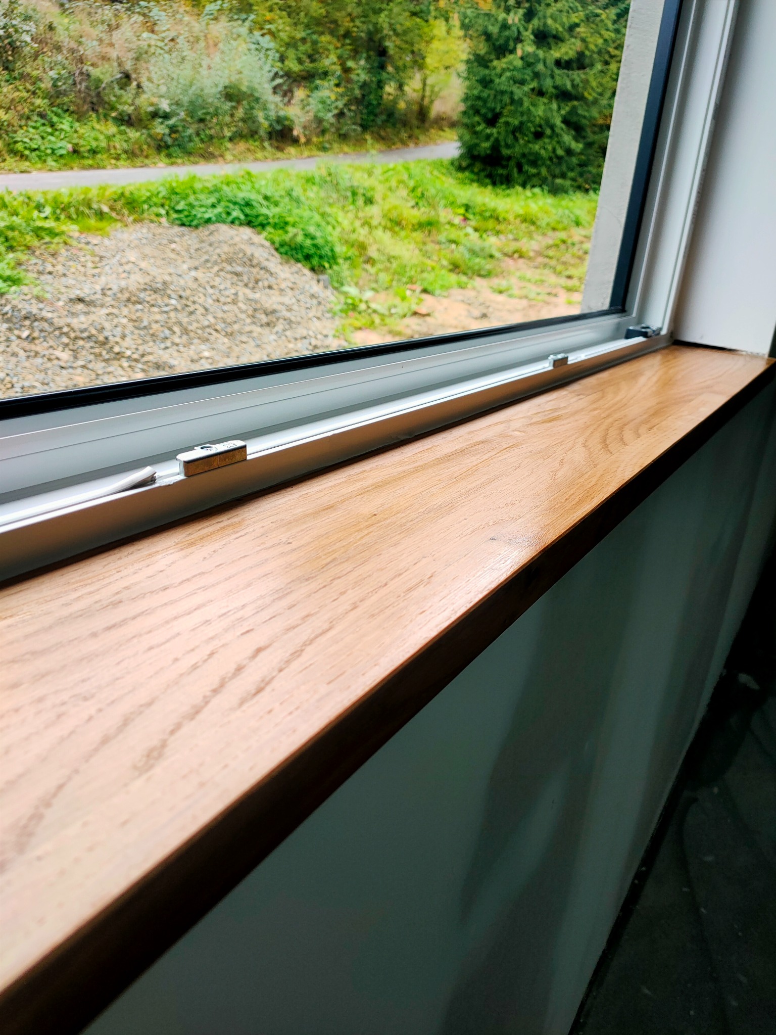 Lakierowany, dębowy parapet wewnętrzny zamontowany w oknie PCV, widok na zewnątrz budynku z zielenią i hałdą żwiru.