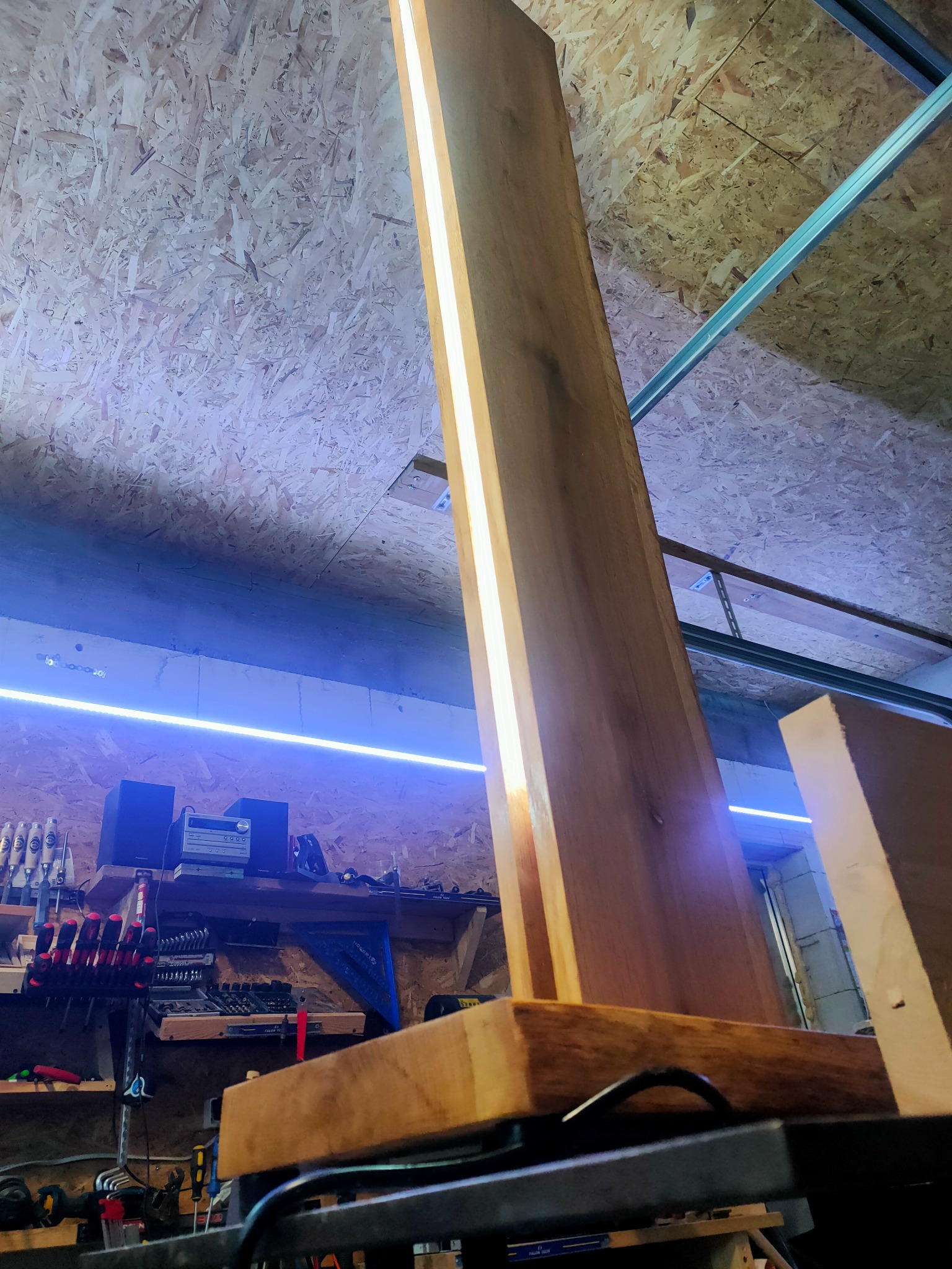 Surowy, wysoki panel drewniany z wbudowanym oświetleniem LED, ustawiony pionowo w warsztacie stolarskim z narzędziami na półkach w tle.