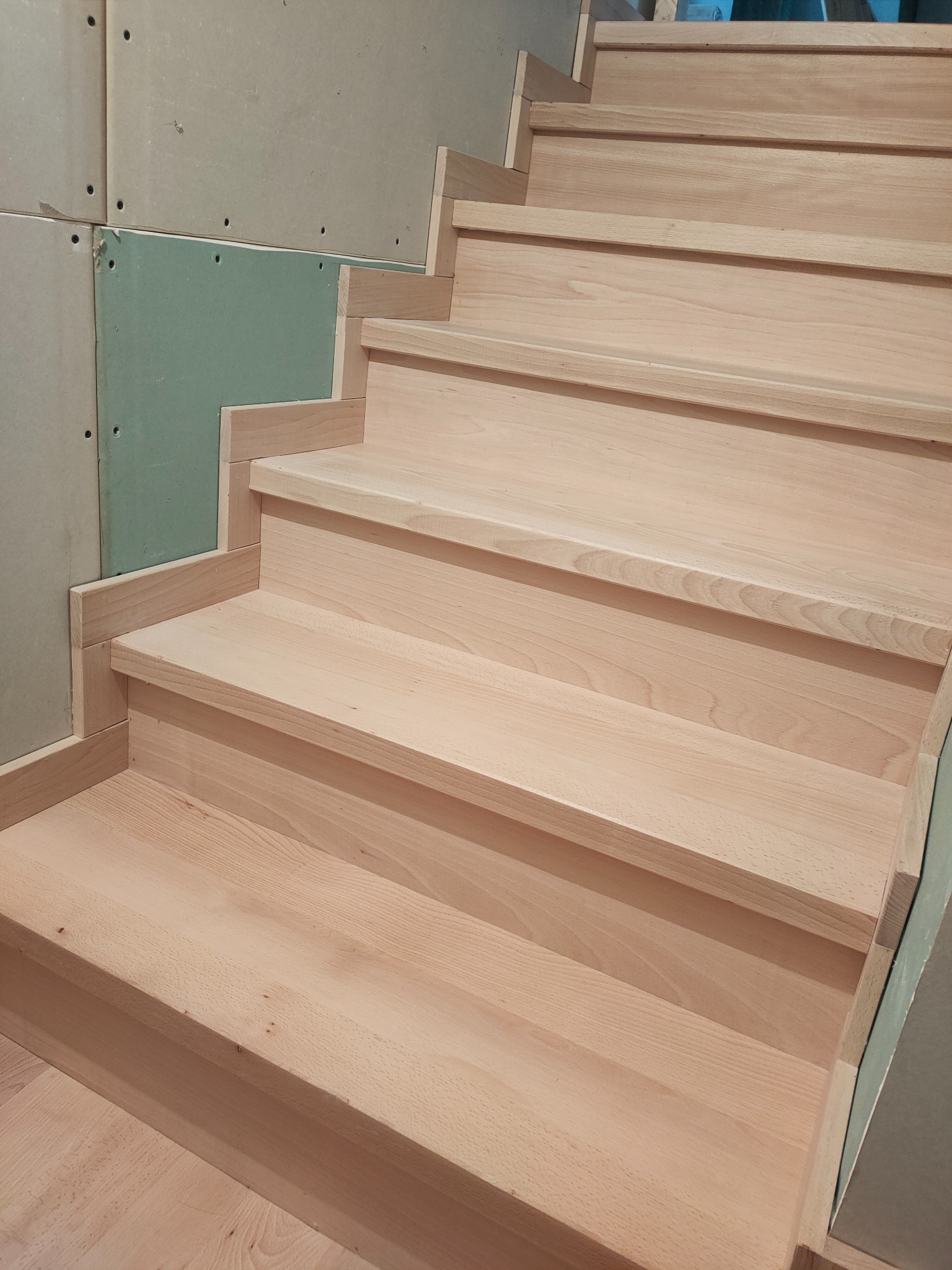 Jasne, drewniane schody wewnętrzne w trakcie montażu, widoczne płyty gipsowo-kartonowe na ścianie, surowe wykończenie.