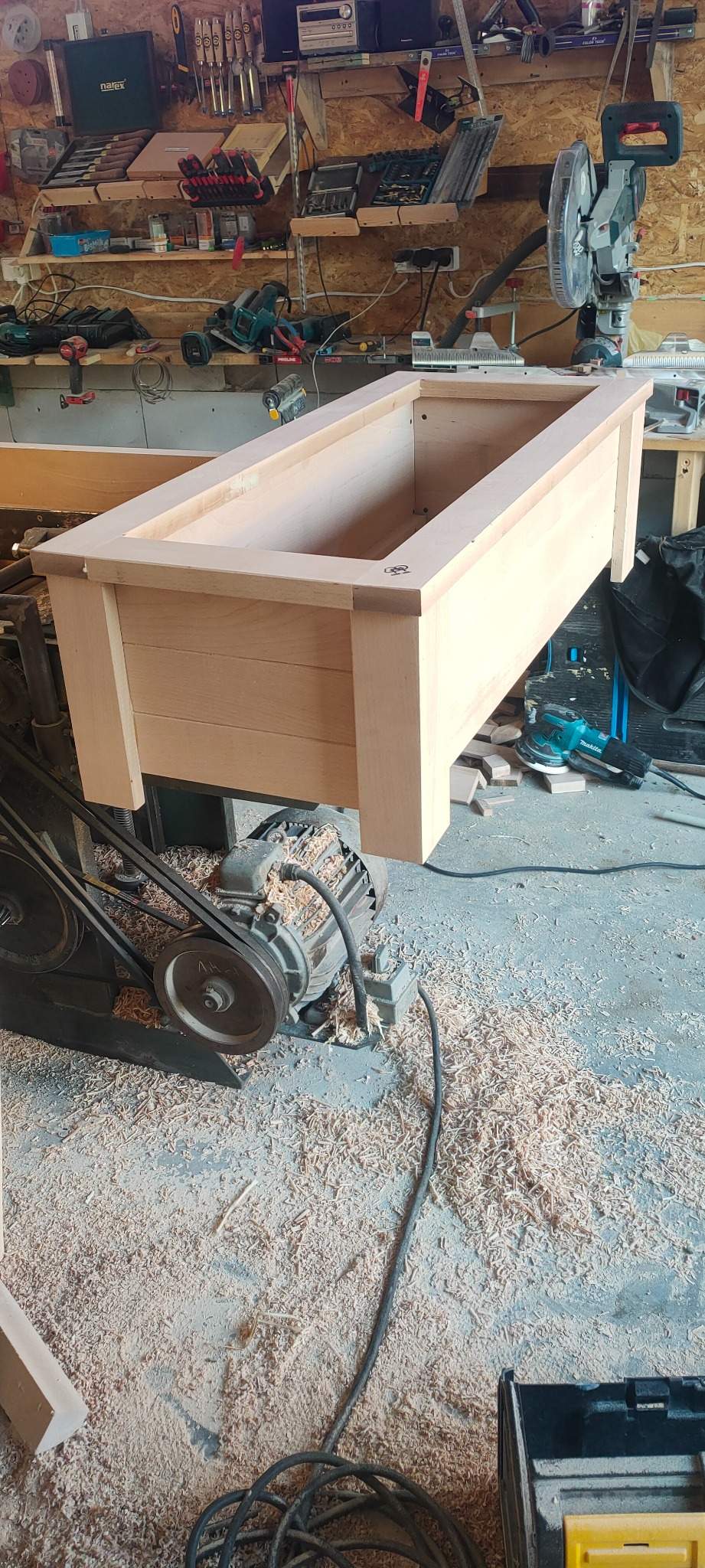 Drewniana skrzynia/doniczka w trakcie tworzenia w warsztacie stolarskim, otoczona wiórami, z widocznym silnikiem obrabiarki i narzędziami na półkach w tle.